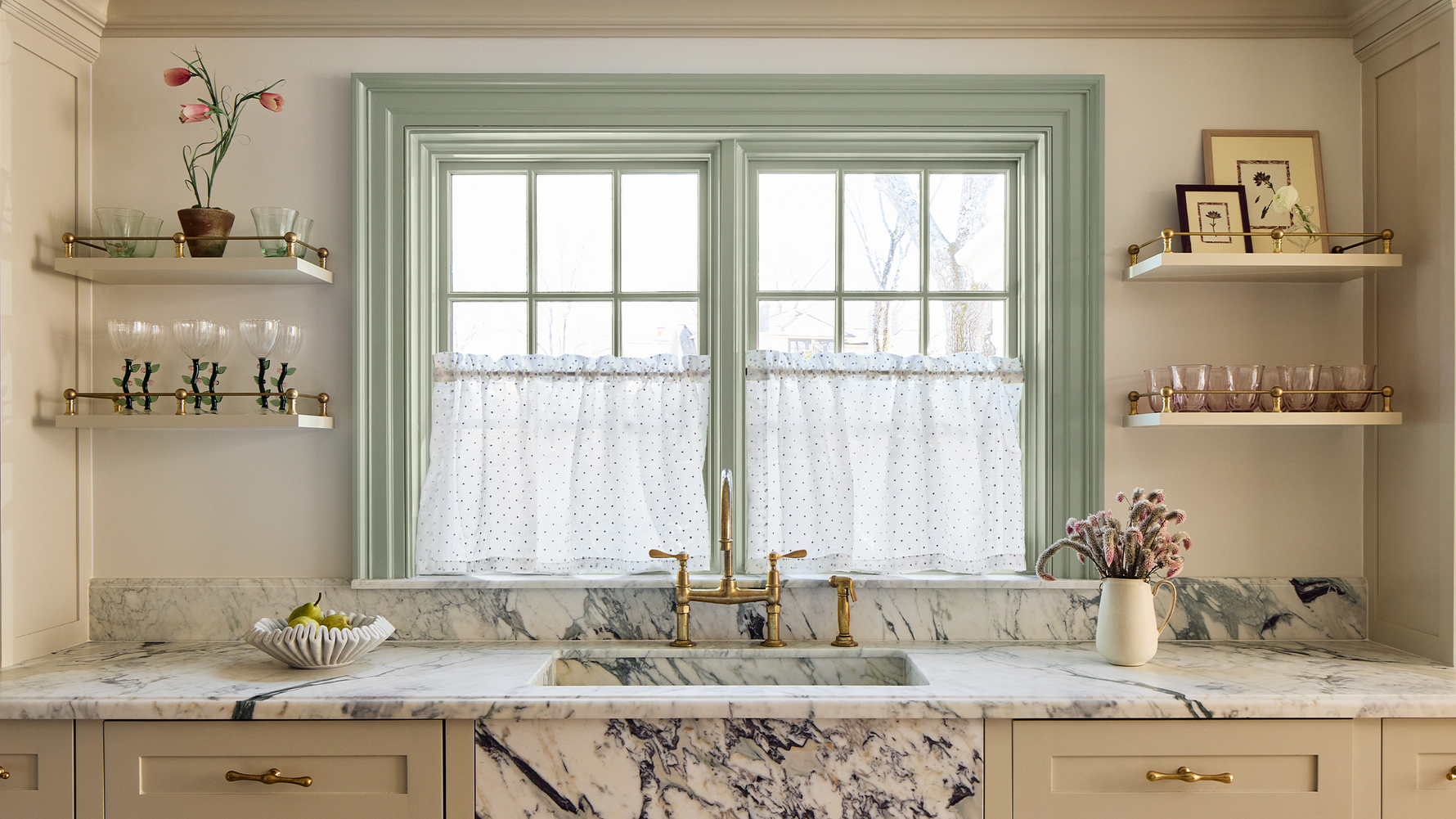 sheer white cafe curtain with embroidered polka dots hung in front of an illuminated window with sage green trim behind a sink in a kitchen with multicolored marble counter and shelves on the wall with glasses and flowers on them