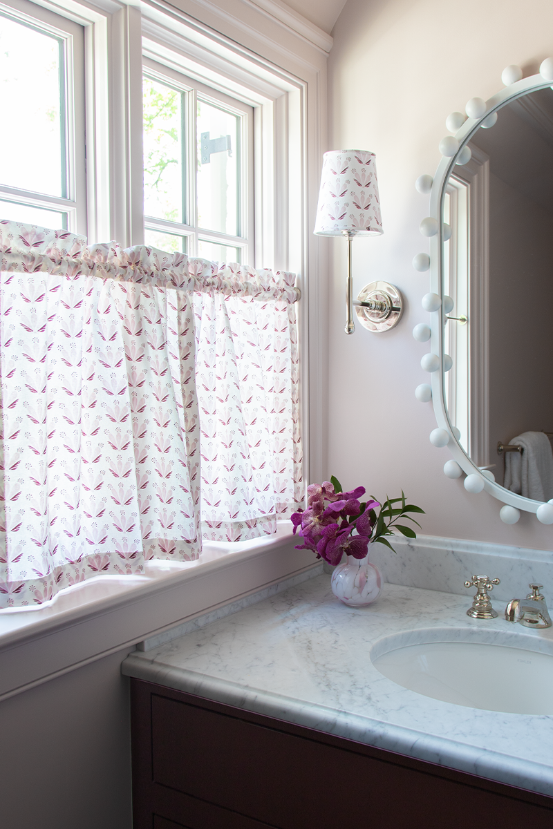 pale pink painted bathroom with white marble counter with a white mirror above the sink and Pink & Burgundy Drop Repeat Floral cafe curtain in front of an illuminated window