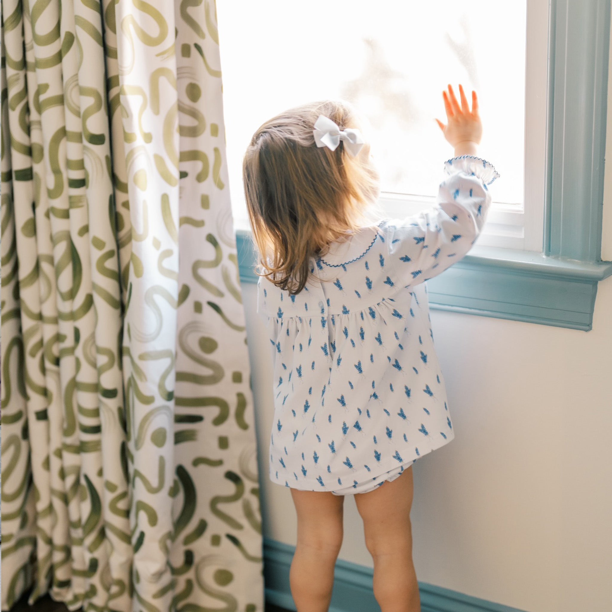 Close-up of Blonde Baby girl with white and blue dress touching the window next to Moss Green Printed Curtain