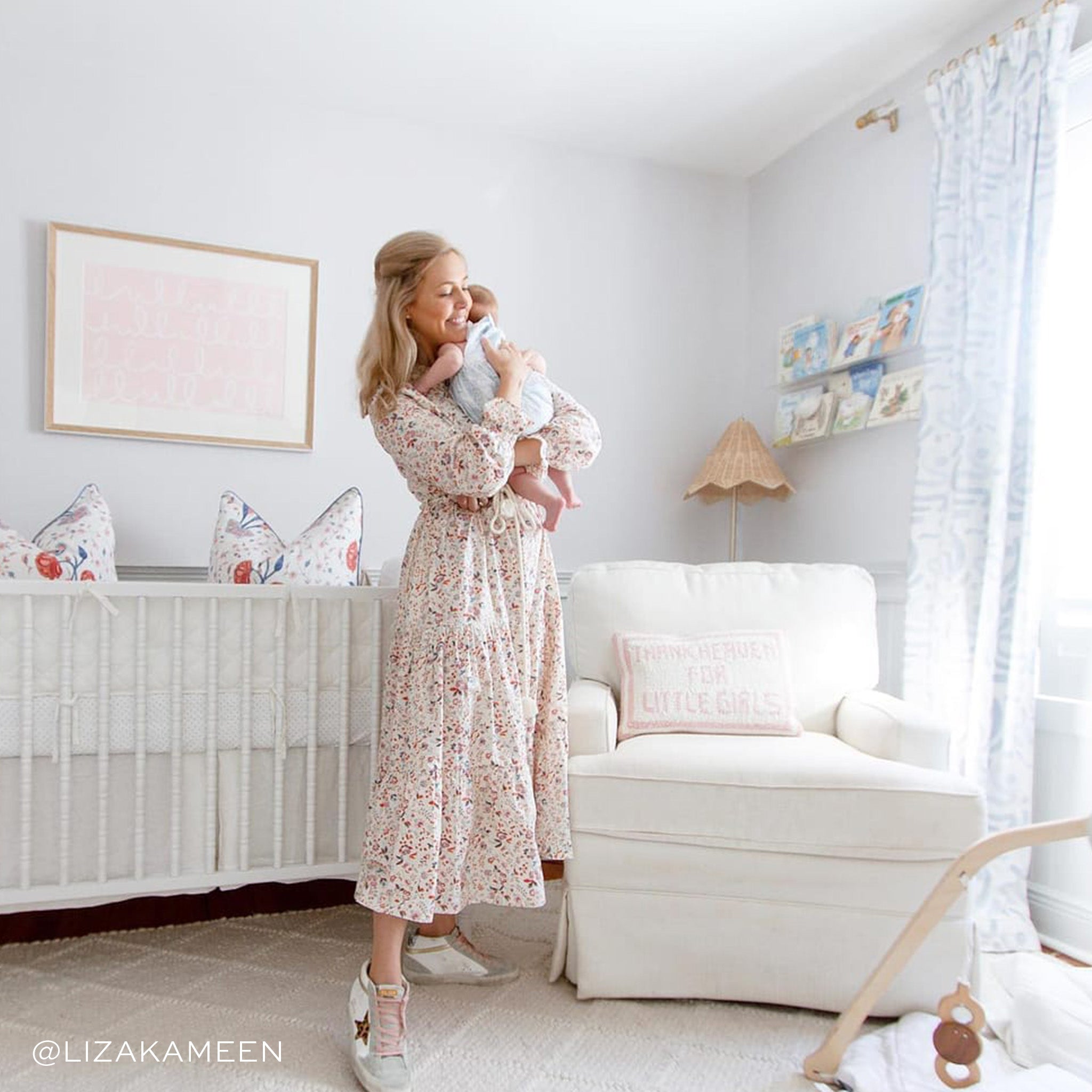 Nursery room with mother wearing floral white dress carrying baby in between a white crib and white couch next to an illuminated window styled with Sky Blue Printed Curtains. Photo taken by Liza Kameen