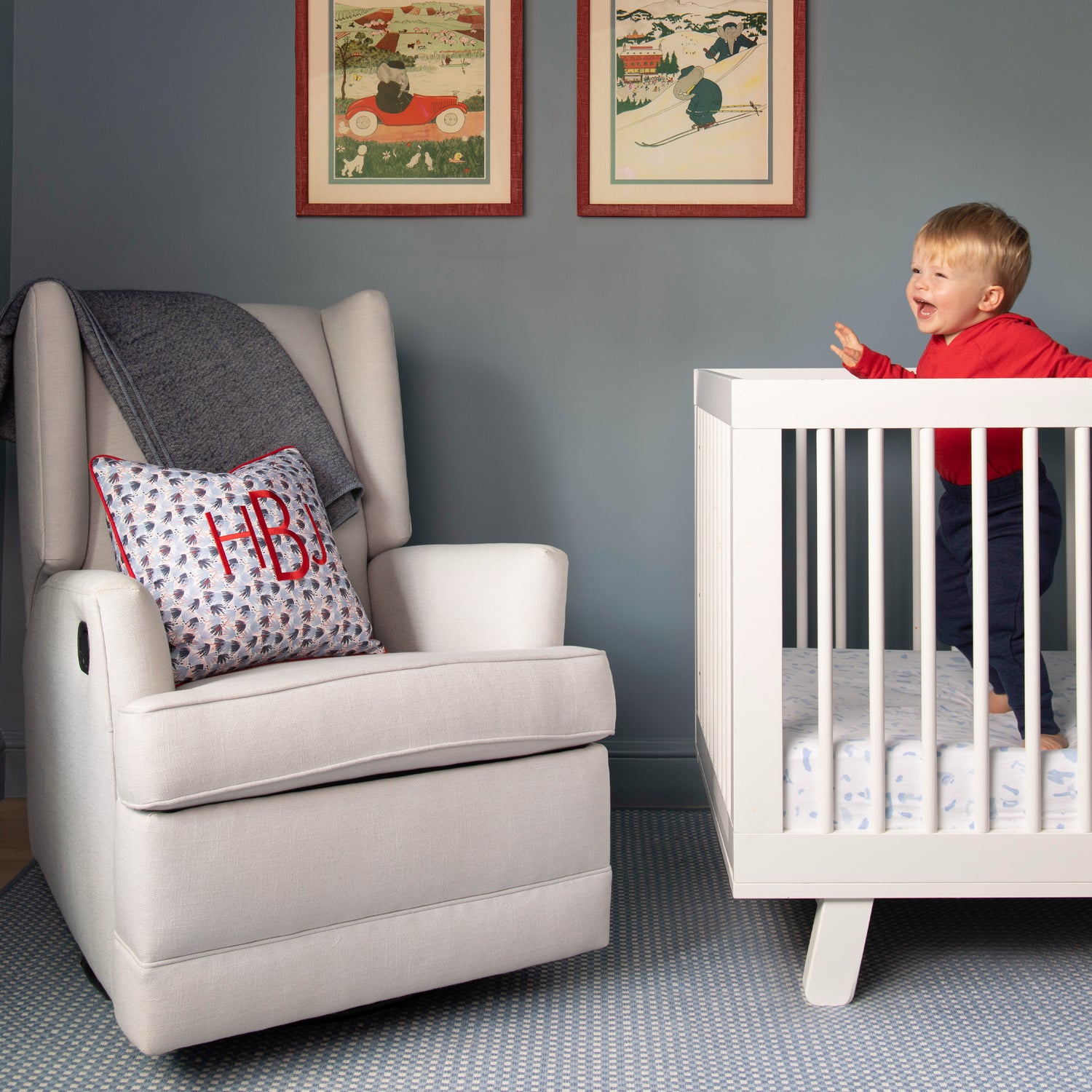 Nursery Room styled with red and blue printed monogrammed pillow on white reclinable sofa chair next to white crib with joyous blonde baby wearing a red long sleeve and navy pants next to two framed artworks