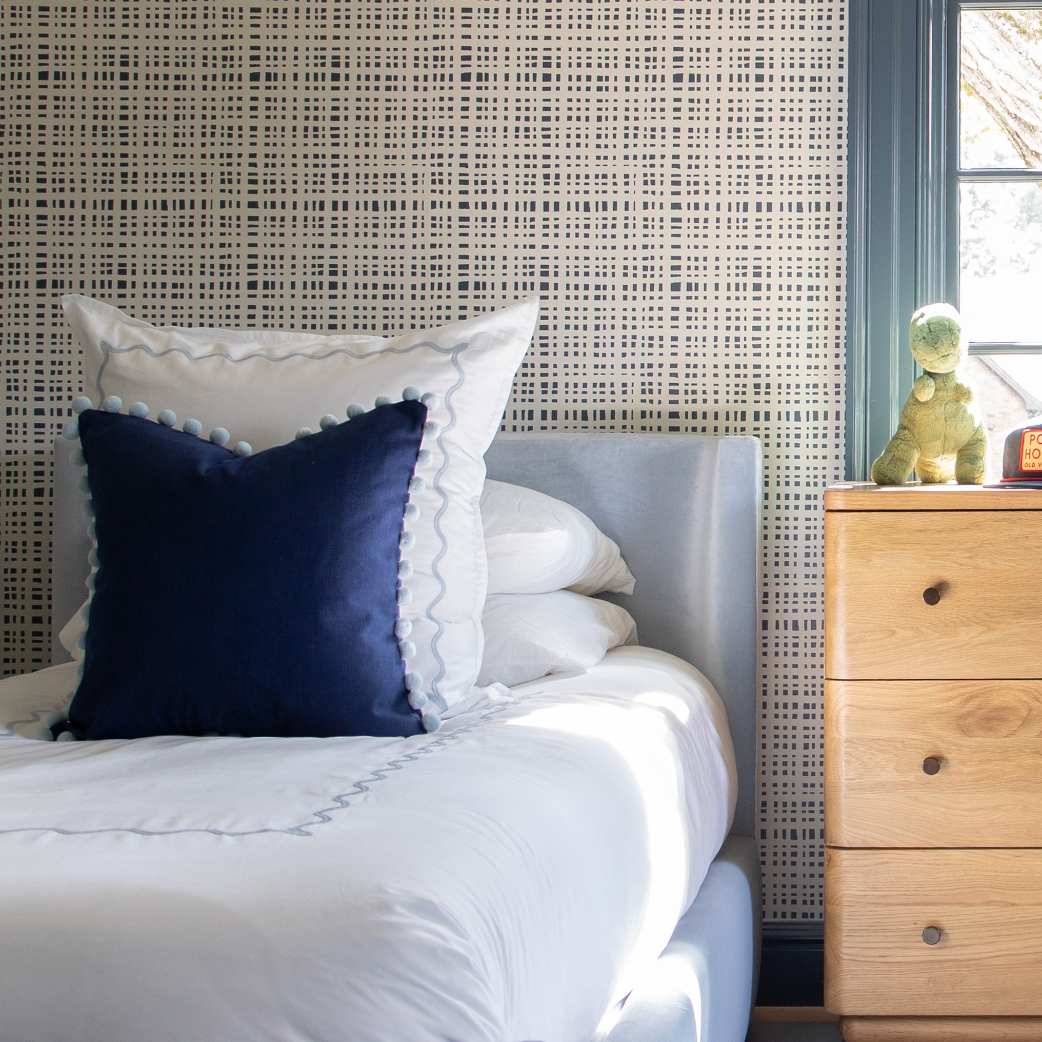 Bedroom styled with navy gingham printed wallpaper and navy blue pillow with sky blue pom pom on white bed by wooden dresser in front of window