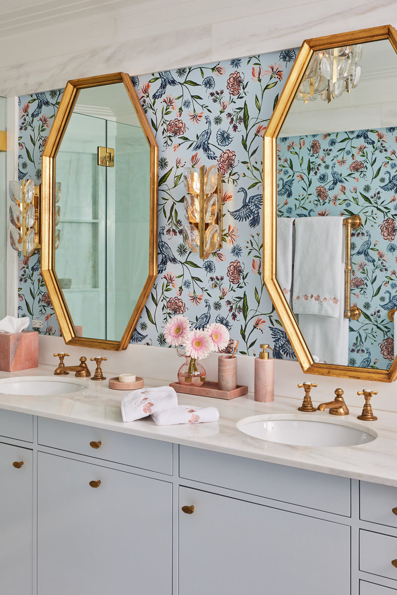 Powder blue chinoiserie wallpaper on a wall in a bathroom with a white marble counter top with two sinks in it with a mirror hung above each sink with gold trim with white towels with pink embroidered botanical stripe stacked on the counter