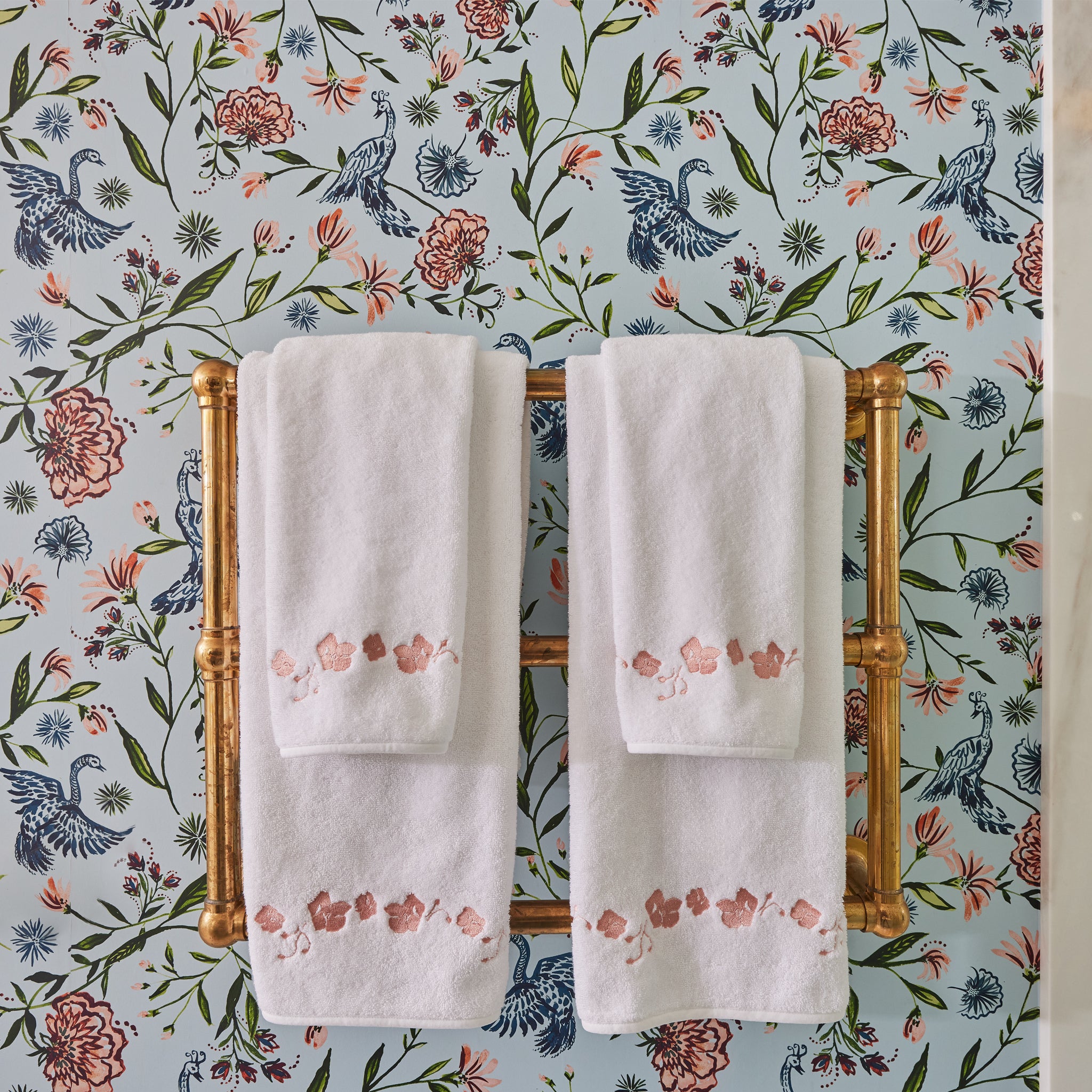 Powder blue chinoiserie wallpaper on a wall in a bathroom with a gold metal towel rack with white towels on it with embroidered coral botanical stripe 