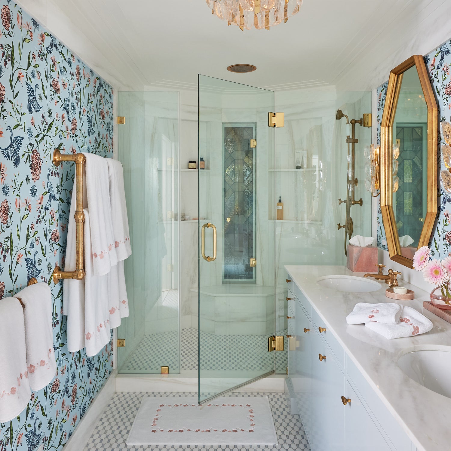 Powder blue chinoiserie wallpaper in a bathroom with white towels with coral embroidered botanical stripe hung on a gold metal rod with a shower and golden trimmed mirrors 