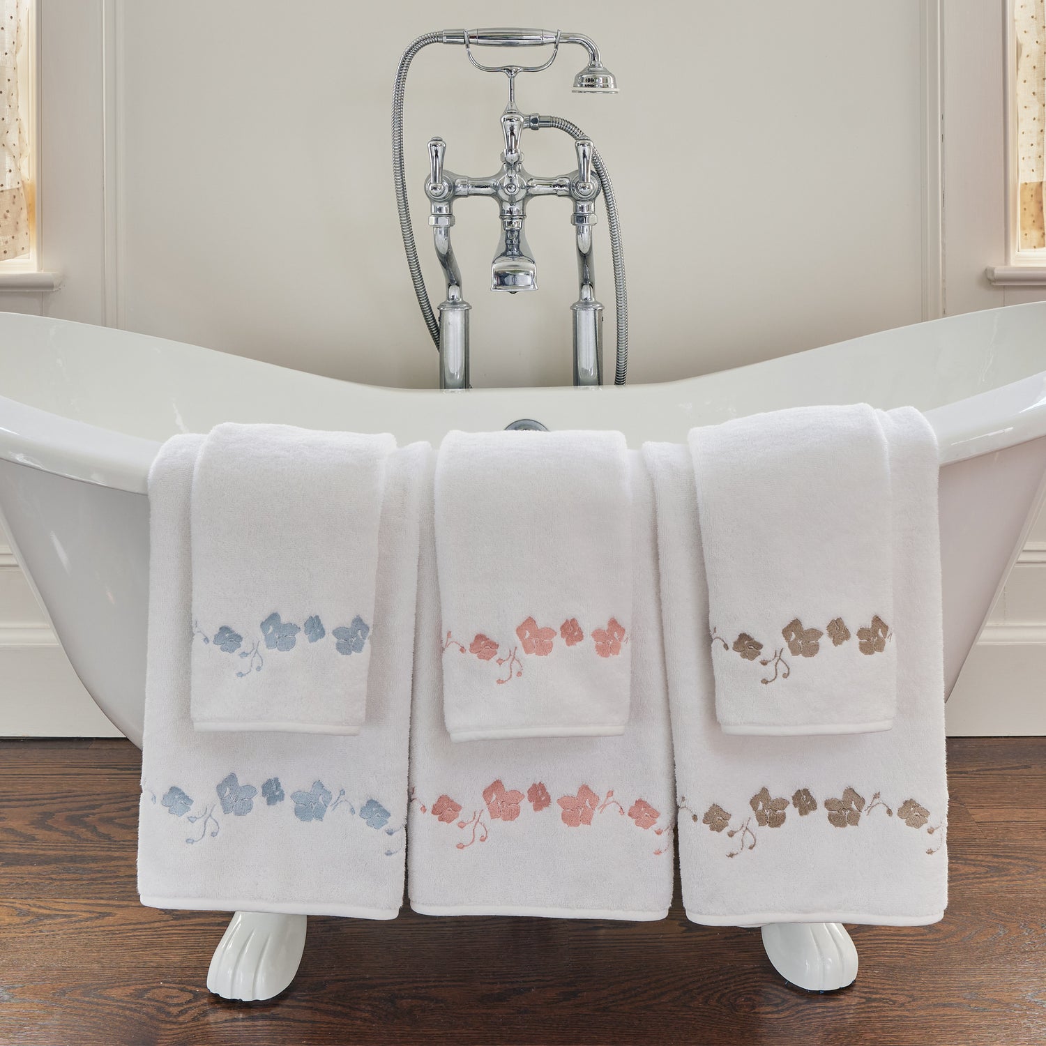 a white bathtub with a set of blue embroidered botanical stripe towels, coral embroidered botanical stripe towels, and sand embroidered botanical stripe pattern hung over the side of the bath tub 