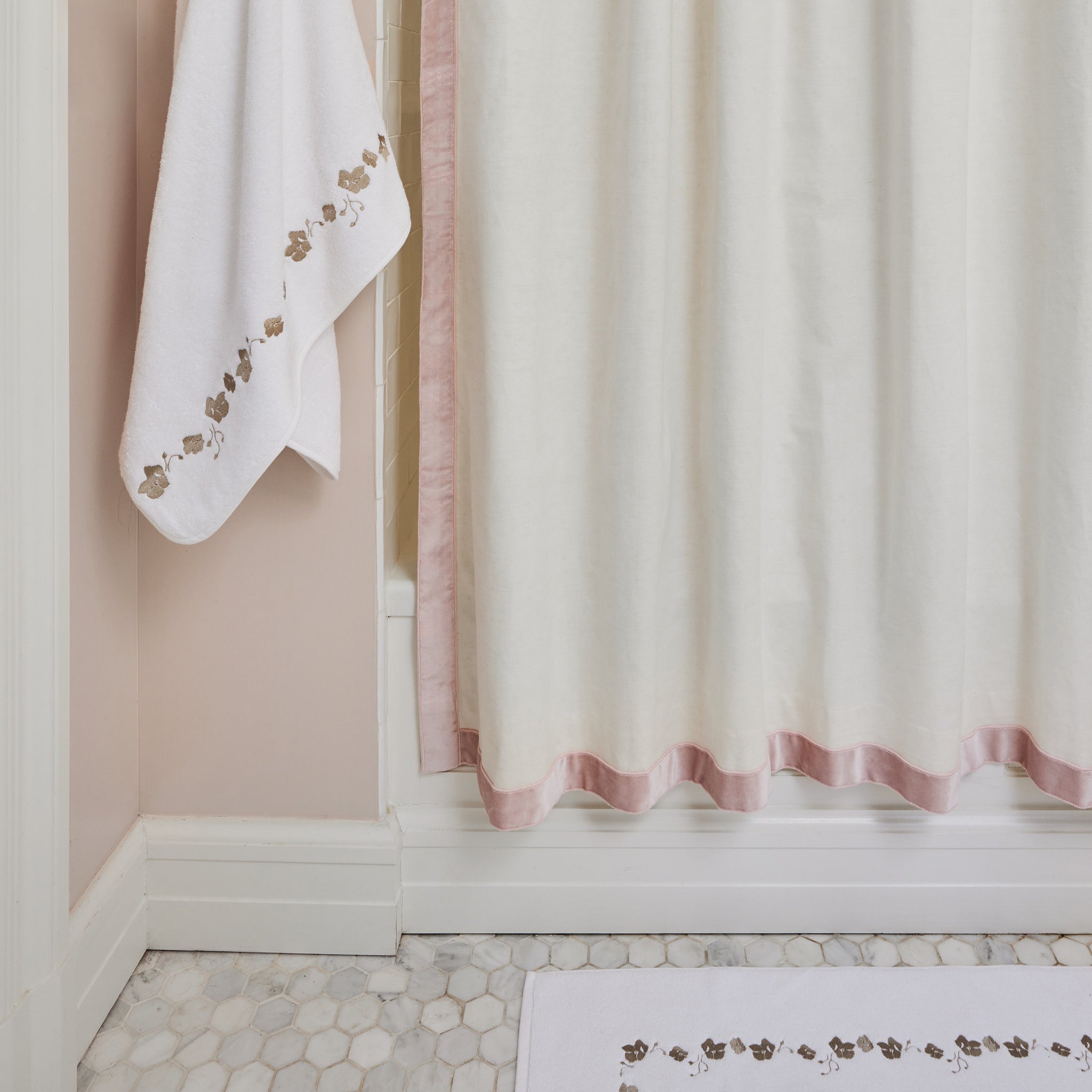 white towel with sand colored embroidered botanical stripe hung next to a shower bath with a white shower curtain with a pink velvet band trim