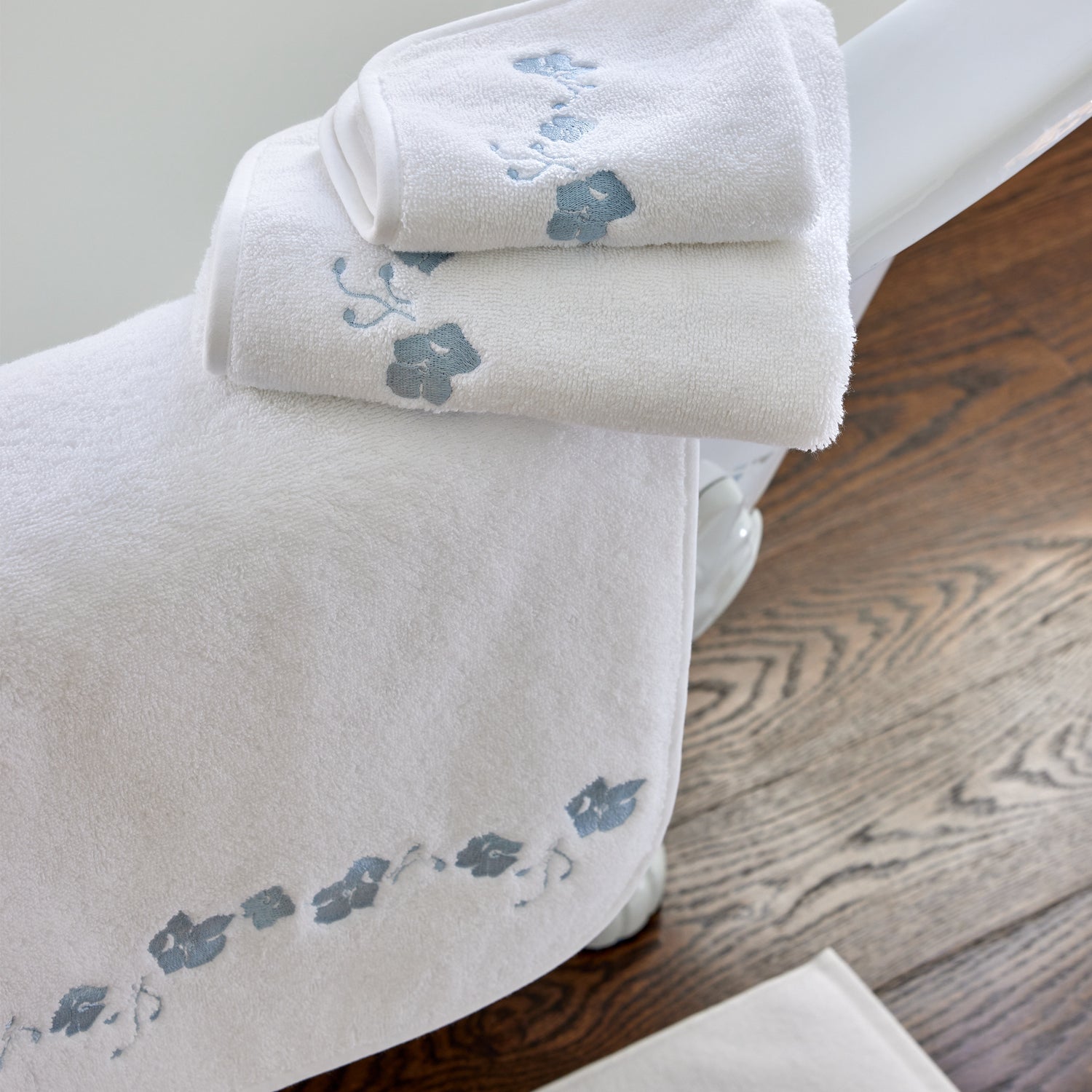 close up picture of a set of white towels with sky blue embroidered botanical stripe on a white bathtub