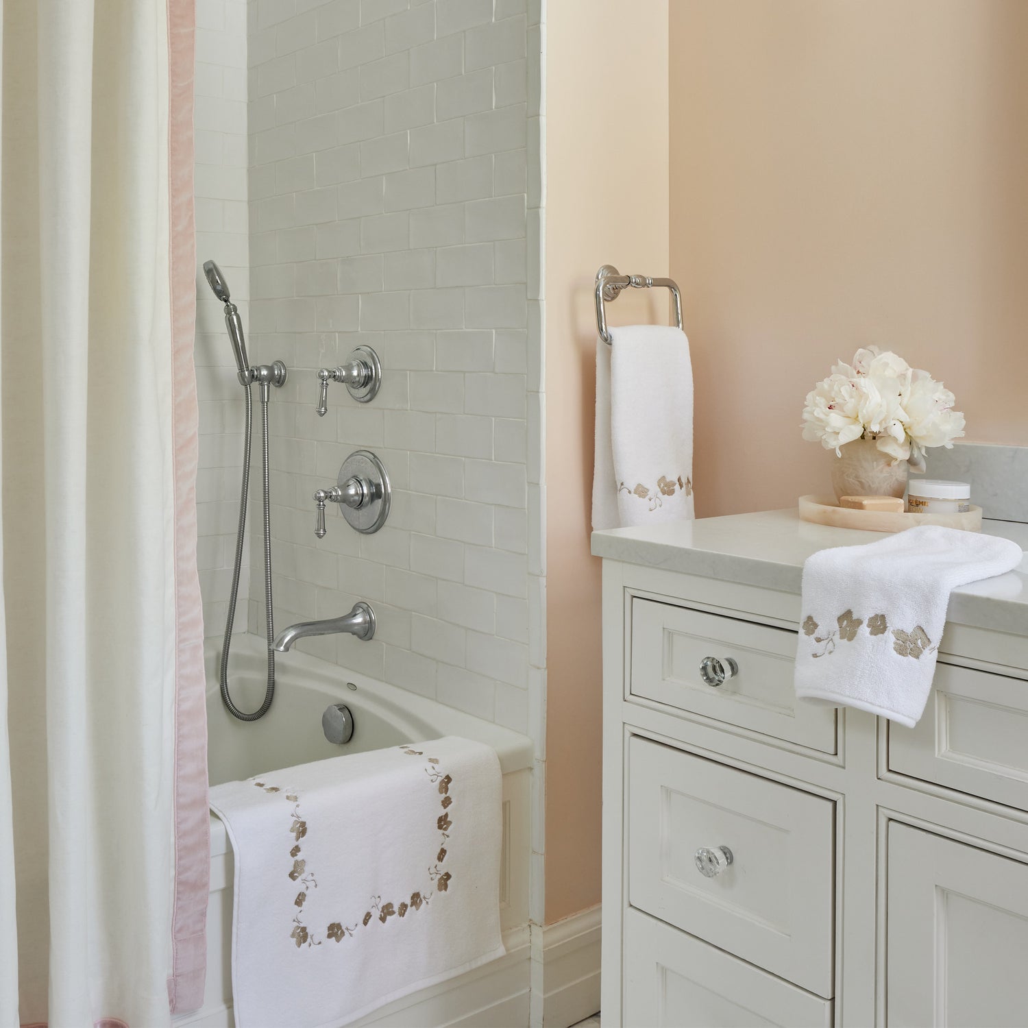 white towel and bath mat with light brown embroidered floral trim in a pink bathroom with a white shower curtain hung with a light pink velvet band and white tiles in the tub