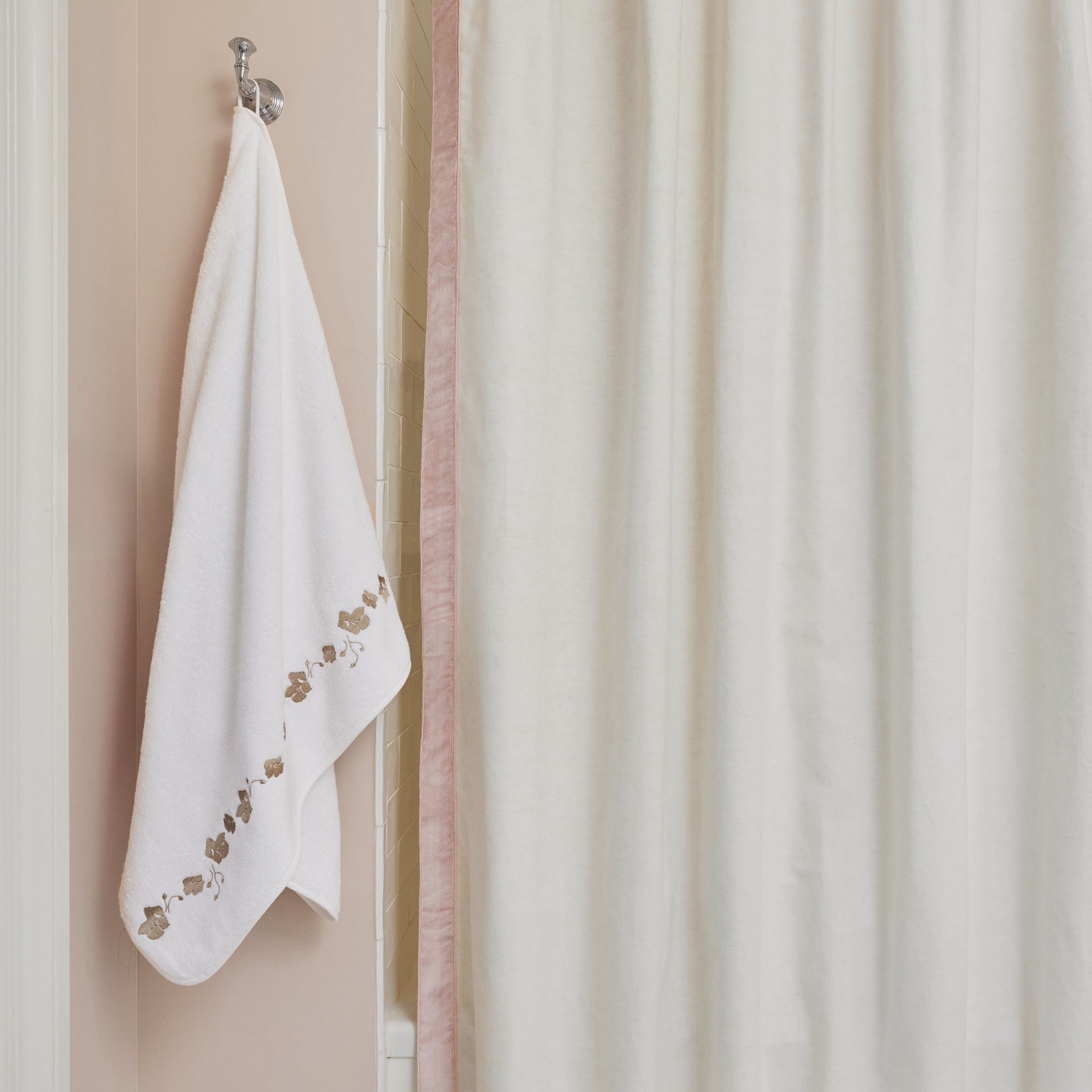 white towel with sand colored embroidered botanical stripe hung on a silver hook next to a shower bath with a white shower curtain with a pink velvet band trim  