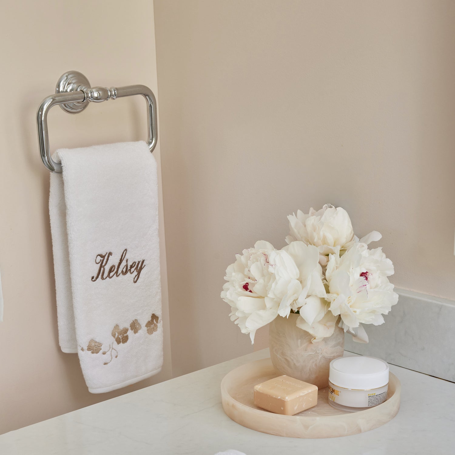 close up of white towel with sand embroidered botanical stripe on a metal towel ring with cream marble bath tray with a bar of soap and a vase of flowers on it sitting on top of a white marble bathroom counter top