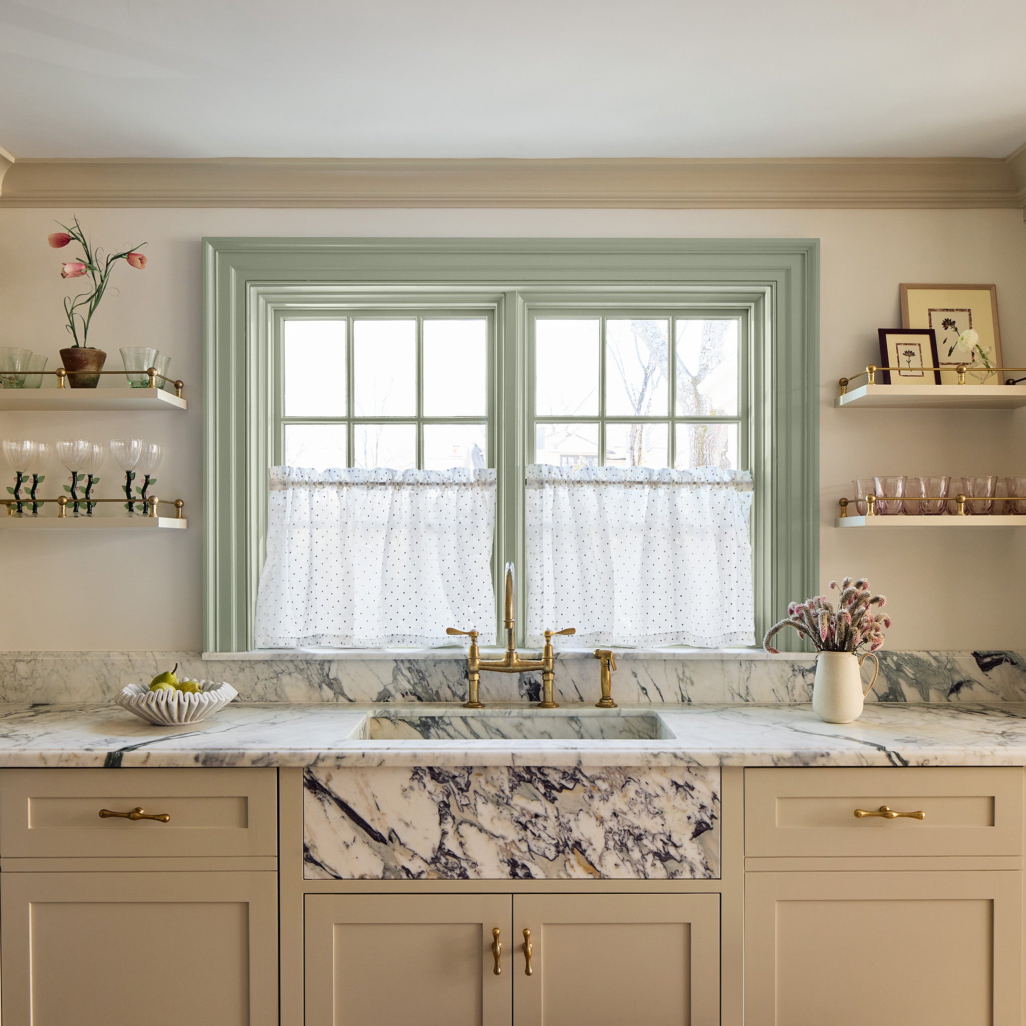 multicolored marble kitchen counter with beige cabinets with gold handles on them, shelves on either side of a window that have glasses on them and framed pictures with sheer embroidered cafe curtains hung in front of an illuminated window with sage green window trim 