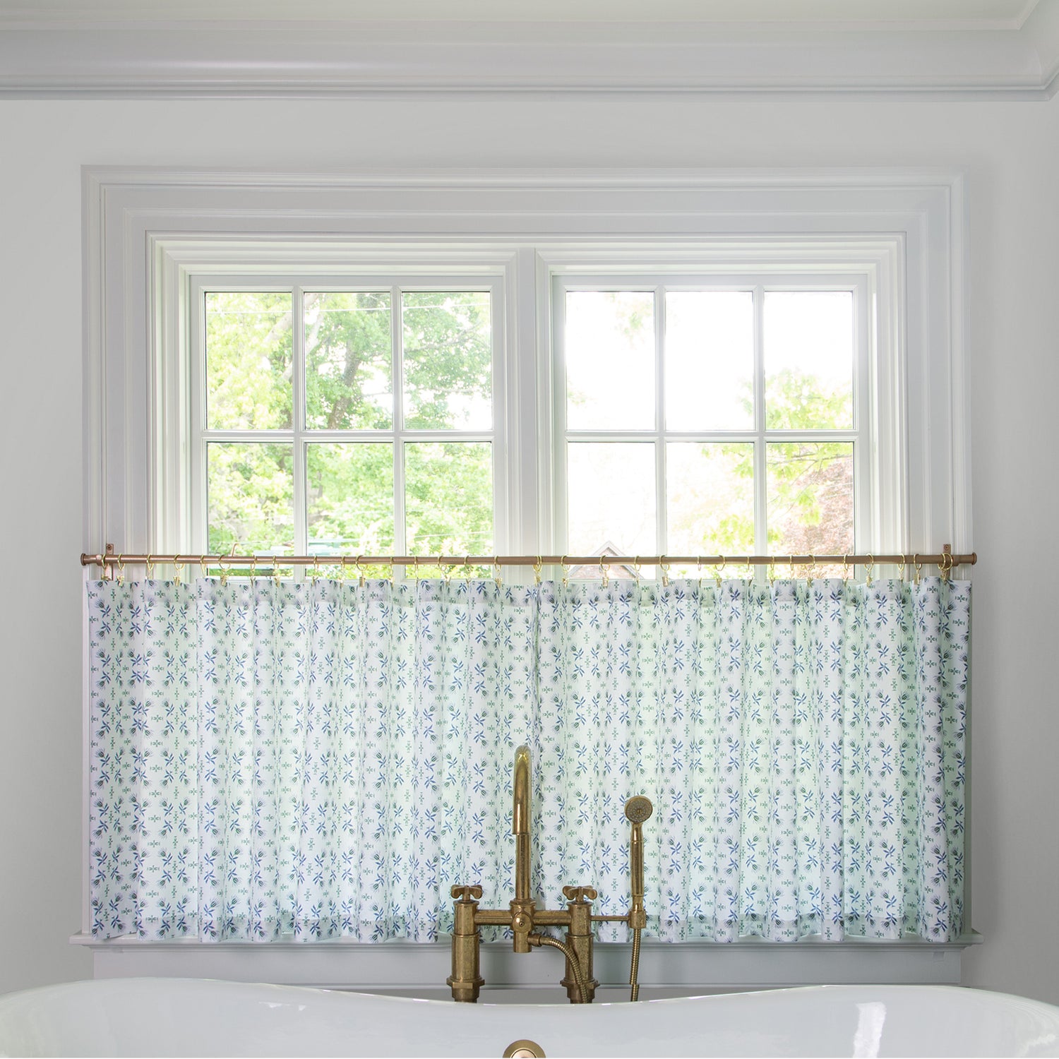 Blue & Green Floral  cafe curtain hung in front of an illuminated window in a bathroom with a white bathtub in front of the window with a gold faucet 