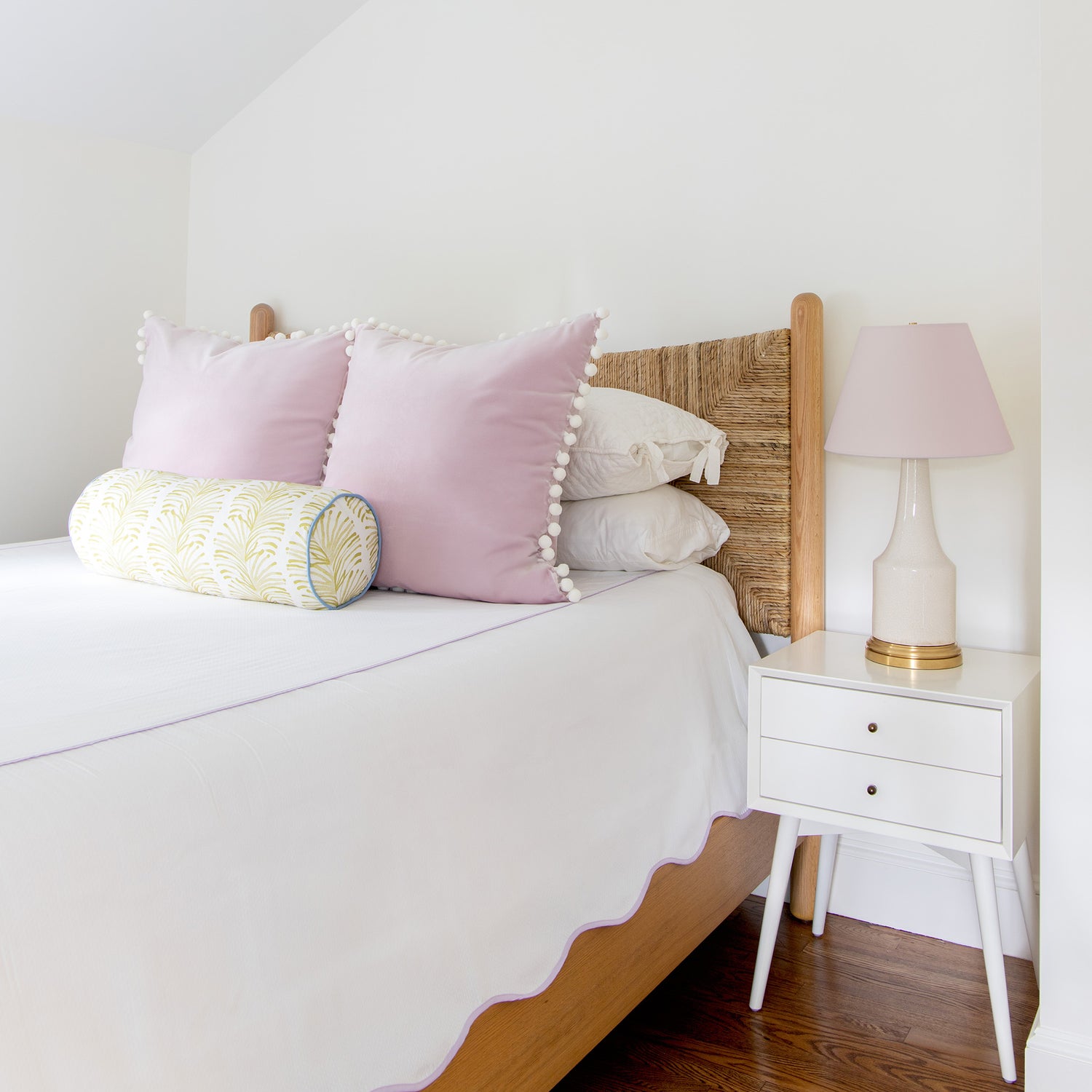 Bedroom styled with two Lilac velvet pillows with white pom poms and Yellow Stripe Chartreuse printed bolster with blue piping on white bed next to white night stand with white, gold, and pink lamp