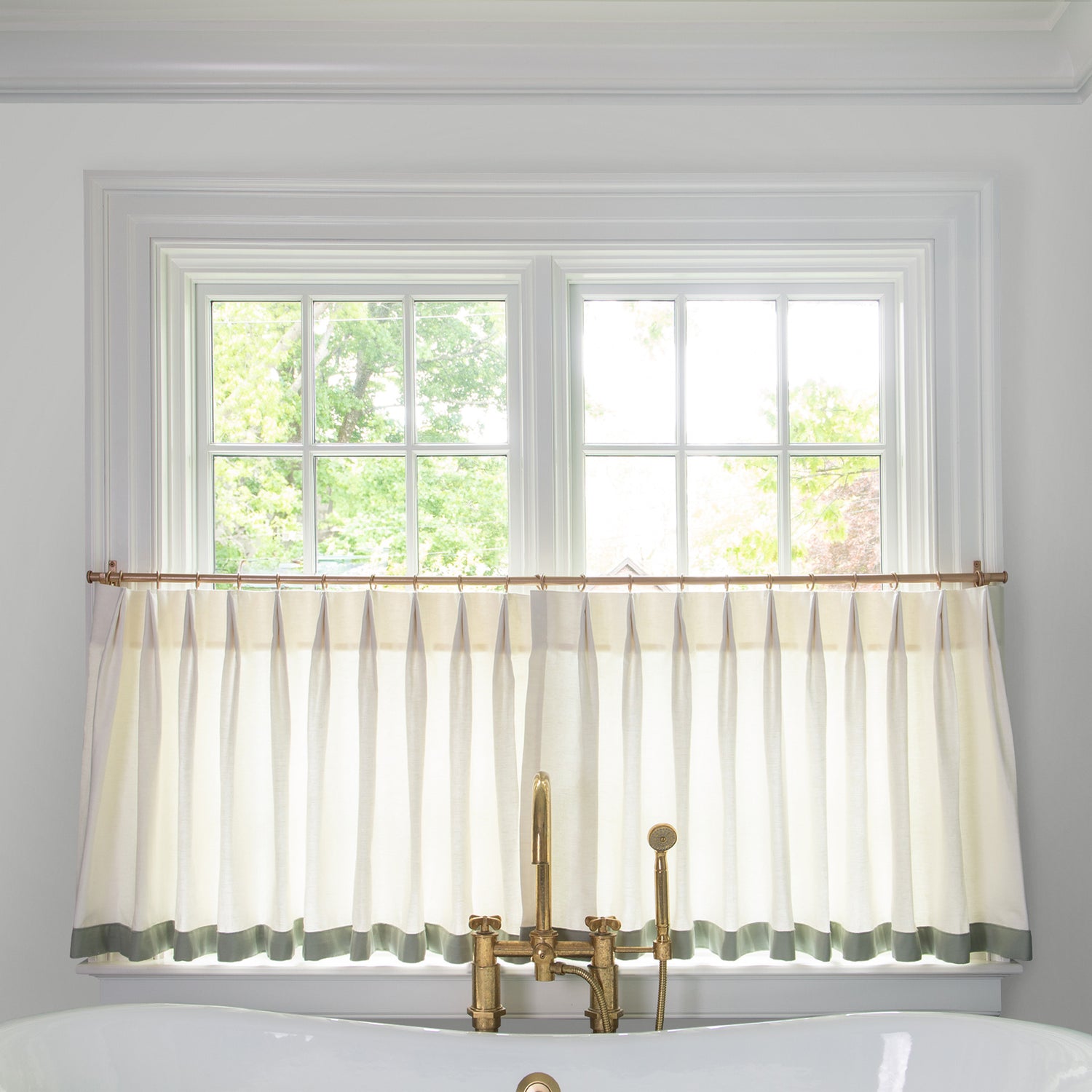  sheer white cafe curtain hung in front of an illuminated window in a bathroom with a white bathtub in front of the window with a gold faucet 