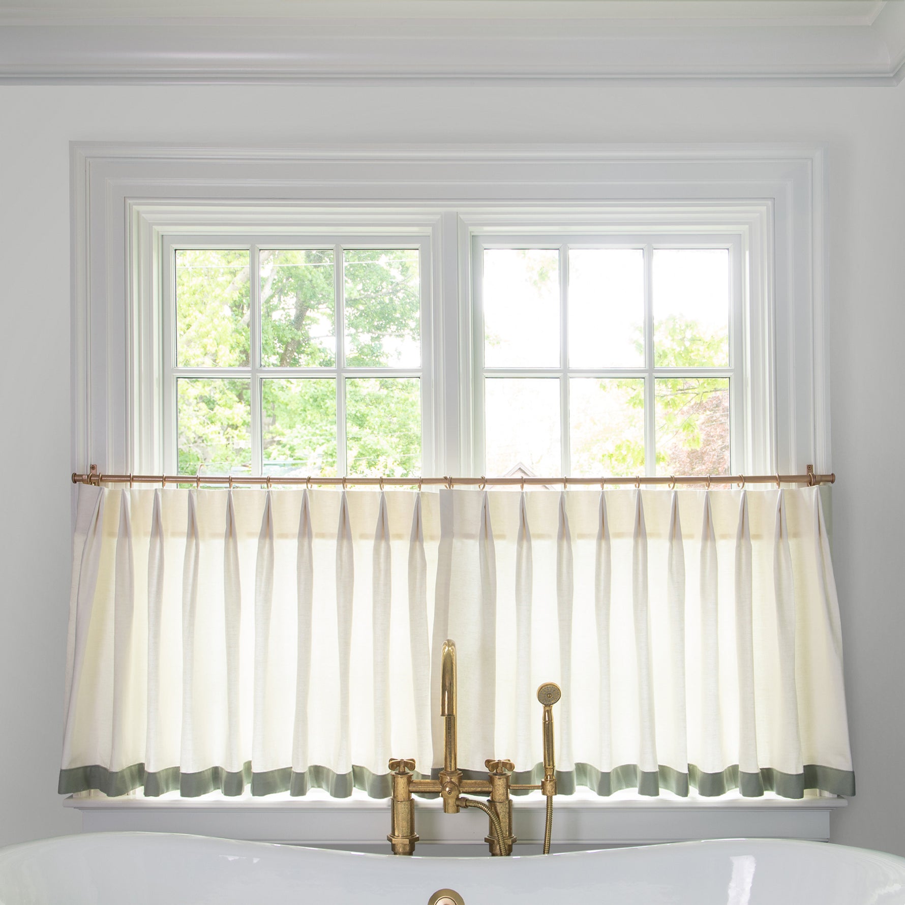  sheer white cafe curtain hung in front of an illuminated window in a bathroom with a white bathtub in front of the window with a gold faucet 