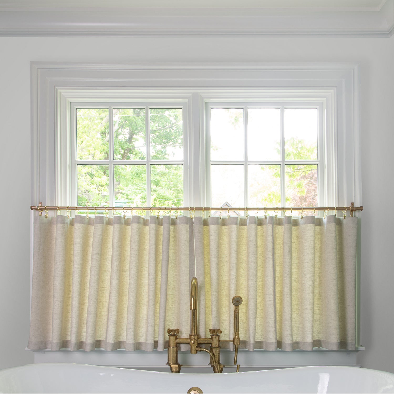 light brown cafe curtain hung in front of an illuminated window in a bathroom with a white bathtub in front of the window with a gold faucet 