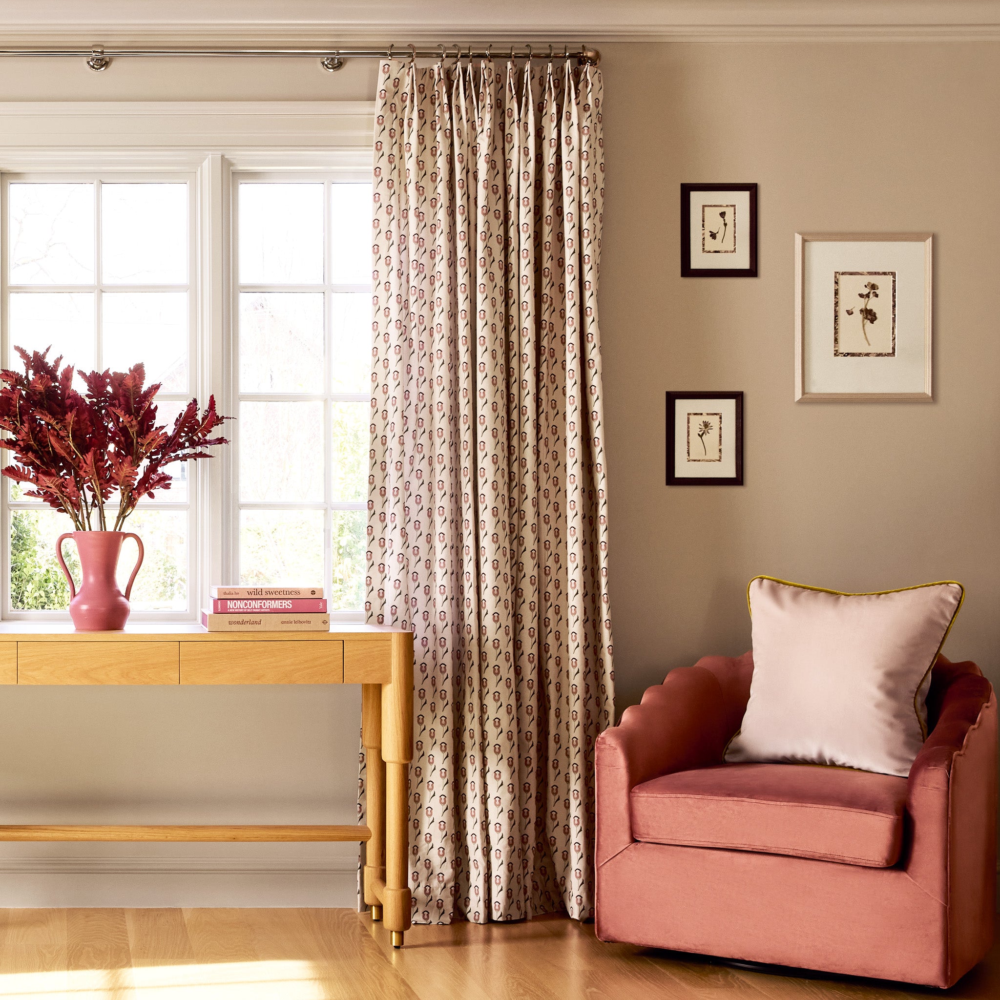 abstract floral pink and green printed curtains on a metal rod in front of an illuminated window with a coral colored chair and botanical artwork on the walls