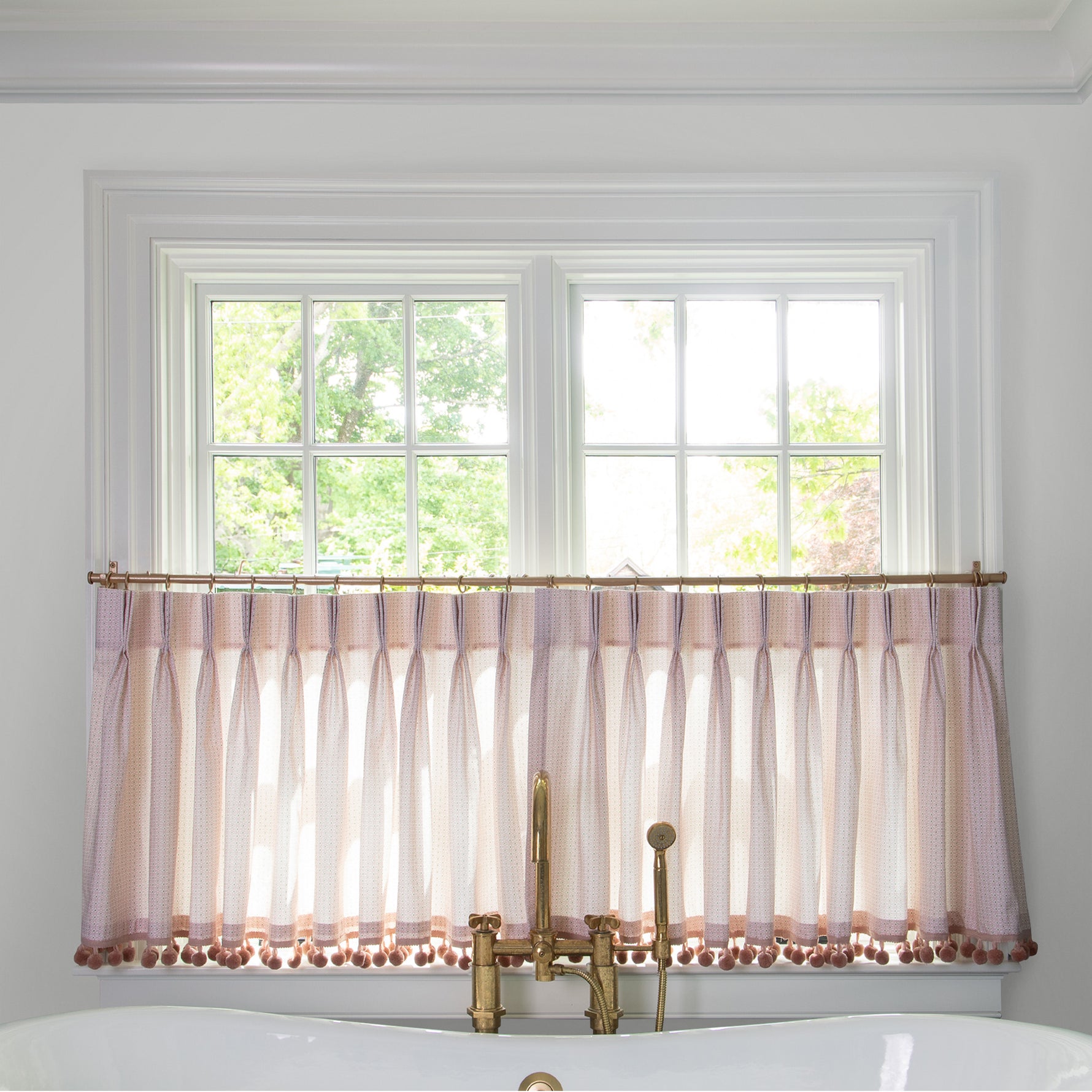 Pink Geometric cafe curtain hung in front of an illuminated window in a bathroom with a white bathtub in front of the window with a gold faucet 