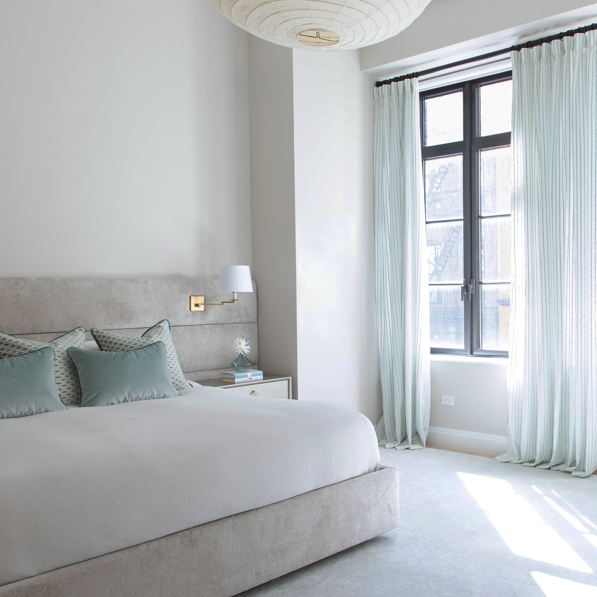 photo of a bedroom with sheer white curtains with mint green embroidered stripes on them hung in front of an illuminated window with a cream velvet bed near the window with white bedding and blue green velvet pillows and mint green geometric chenille jacquard pillows on the bed 