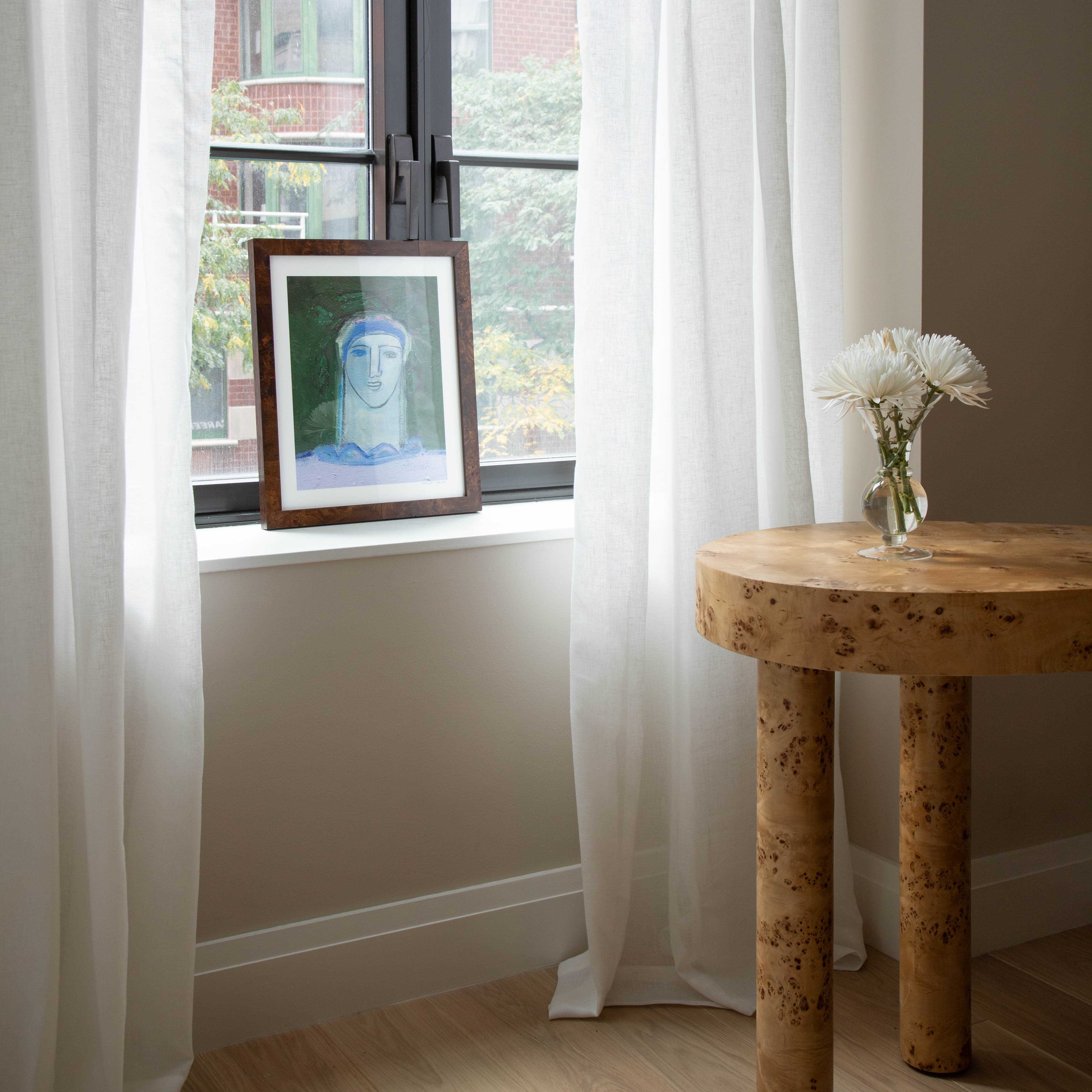 sheer white curtains hung in front of a window with an abstract painting of a face sitting on the window sill and a light brown wooden table with a clear vase with white flowers in the vase