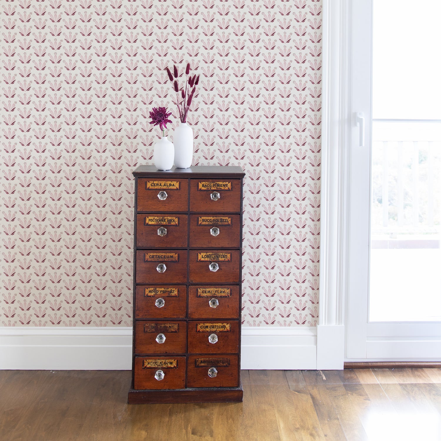 Corner close-up styled with a Pink & Burgundy Drop Repeat Floral Printed Wallpaper behind wooden cabinet with two white vases with pink flowers inside