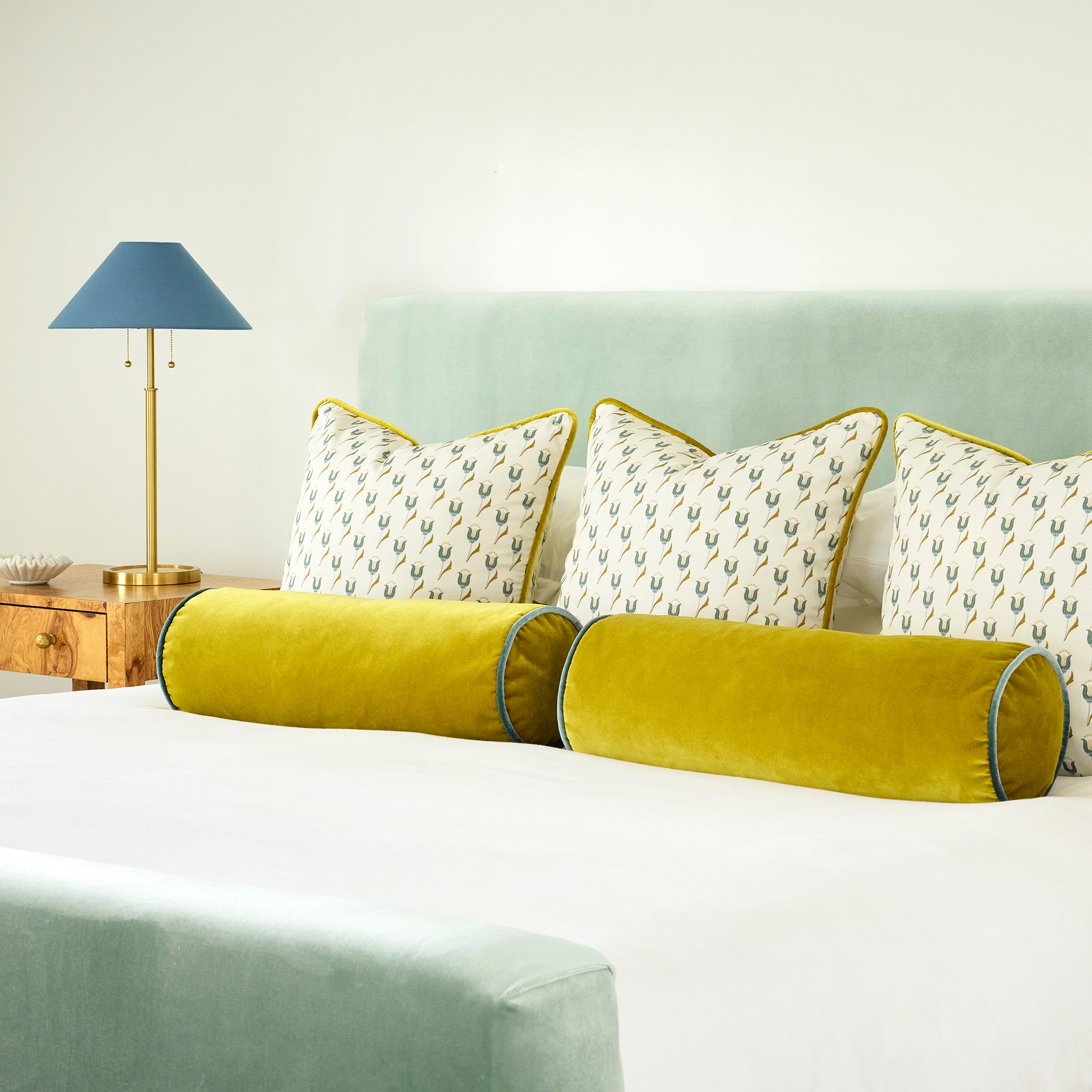 Light blue green bed frame with white bedding, abstract floral green pillows, and and gold yellow bolster pillows on the bed, a wooden night stand next to the bed with a golden lamp with a blue lampshade on the table