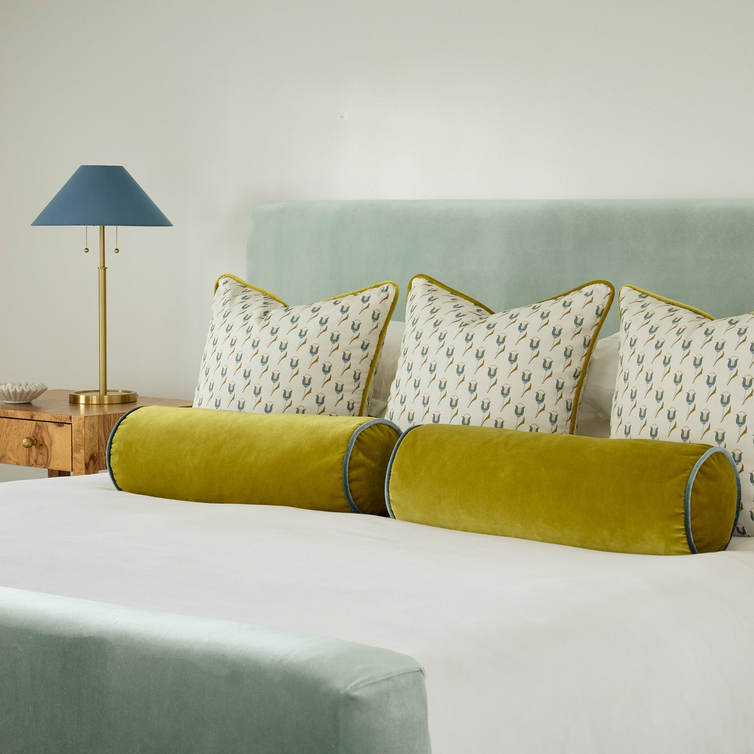 Abstract floral blue and green pillows and golden chartreuse pillows on a bed with white bedding and a brown wooden night table with a gold and blue lamp