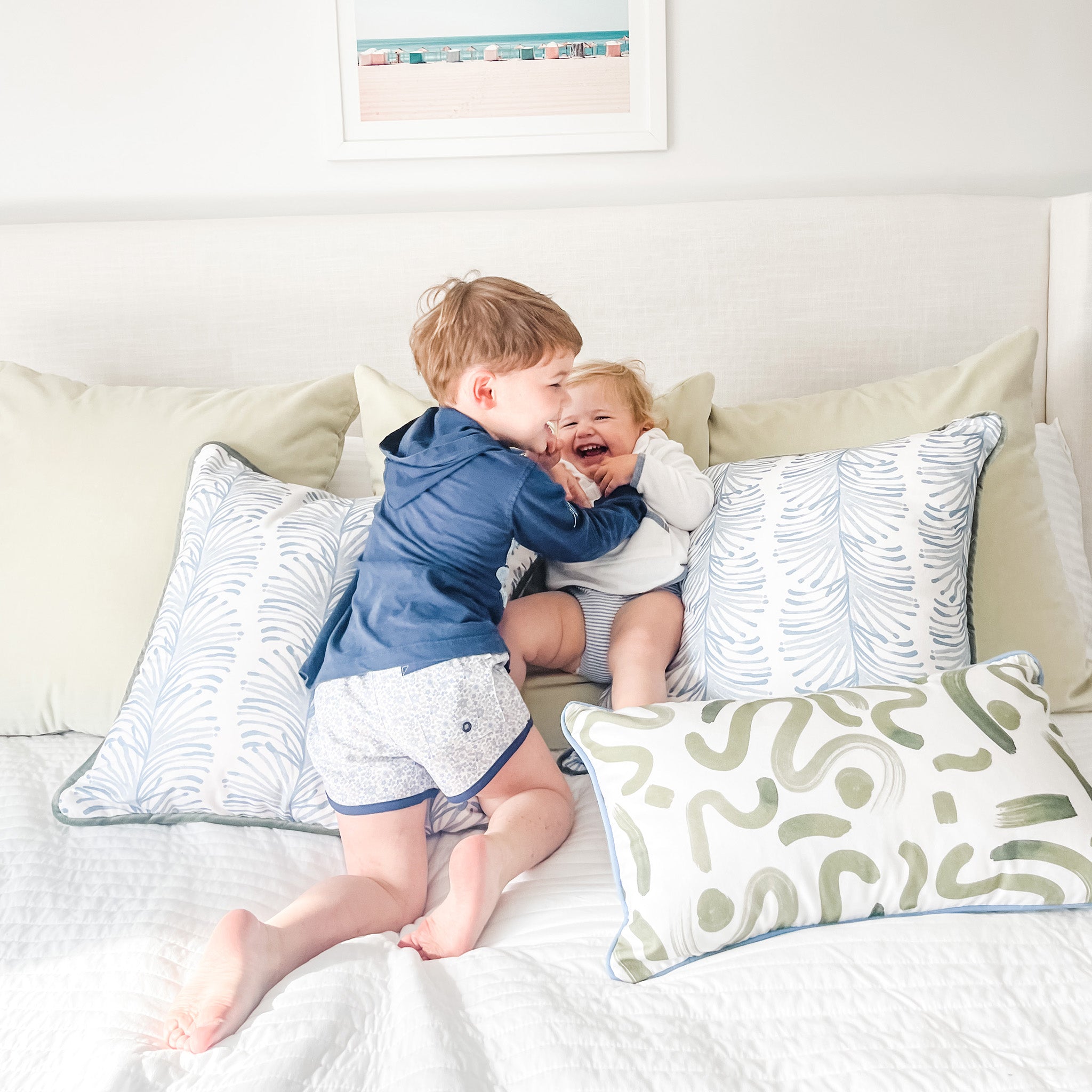 Close-up of bed styled with three light green velvet pillows, two sky blue botanical stripe printed pillows, and one Moss Green Printed Lumbar under two children laughing while playing