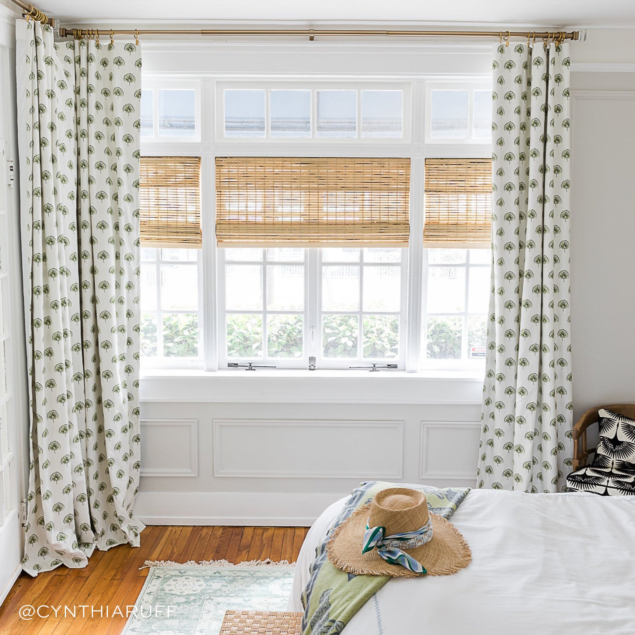 Illuminated window styled with Green Floral Printed Curtain next to white bed with beach hat on top. Photo taken by Cynthia Ruff