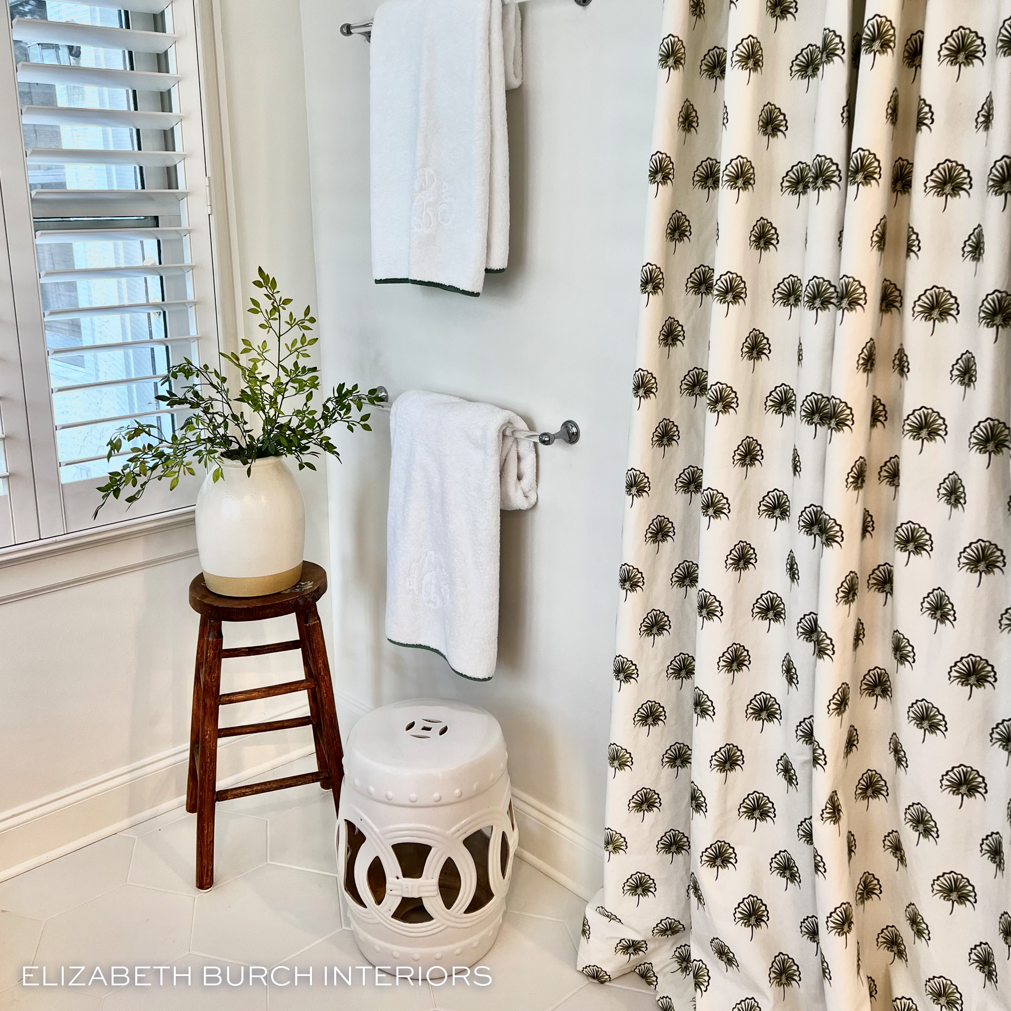 Green Floral Printed shower curtain hanging in front of a white plant stand, white towels and a green plant on a brown stool