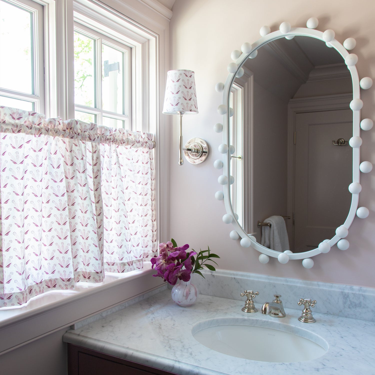 pale pink painted bathroom with a white marble counter top and a white mirror hung above the sink, Pink & Burgundy Drop Repeat Floral cafe curtains in front of an illuminated window and Pink & Burgundy Drop Repeat Floral fabric on a lampshade 