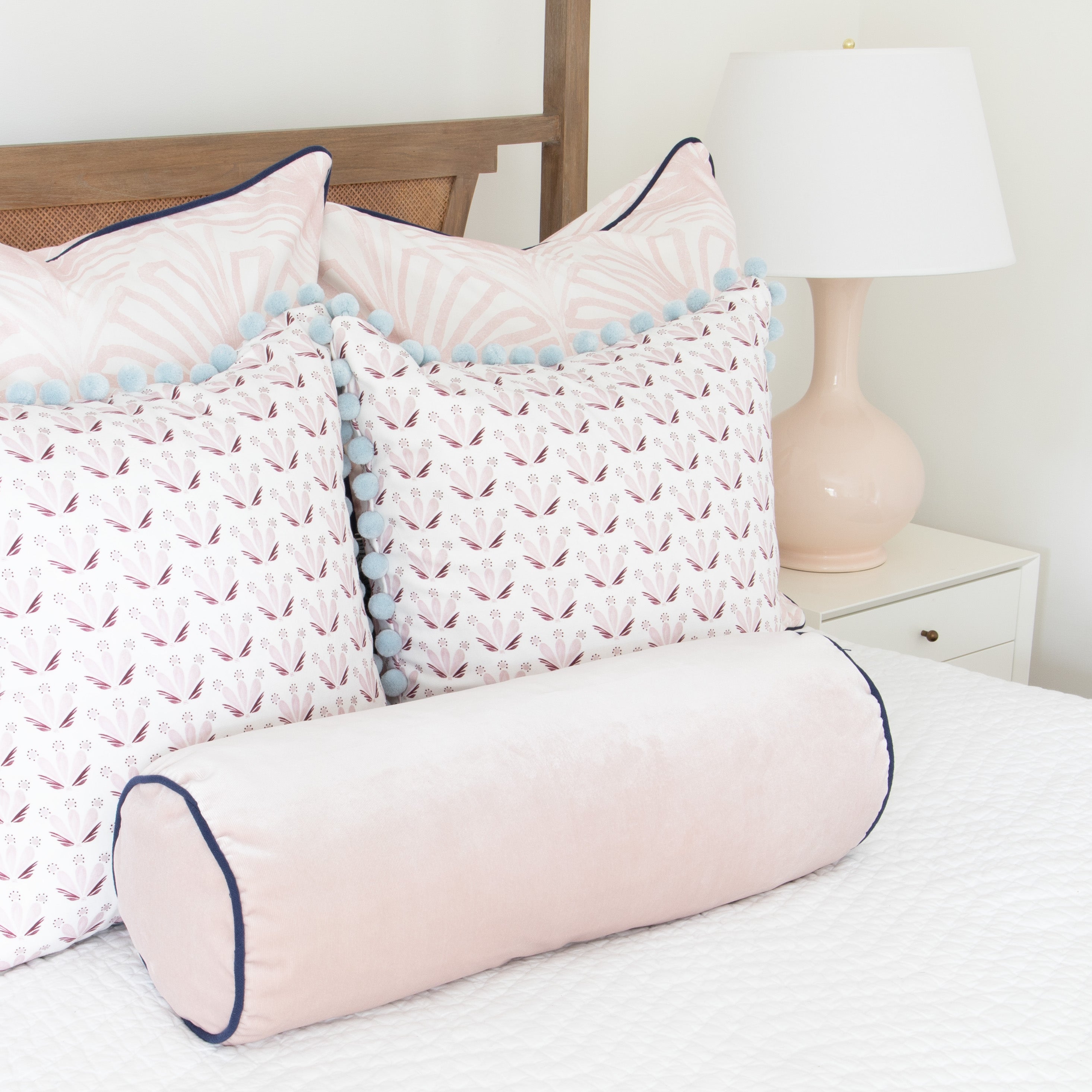 close up of bed with white bedding, pink palm pillows, pink and burgundy floral drop repeat pillows with blue pom poms, and a pink velvet bolster pillow on the bed