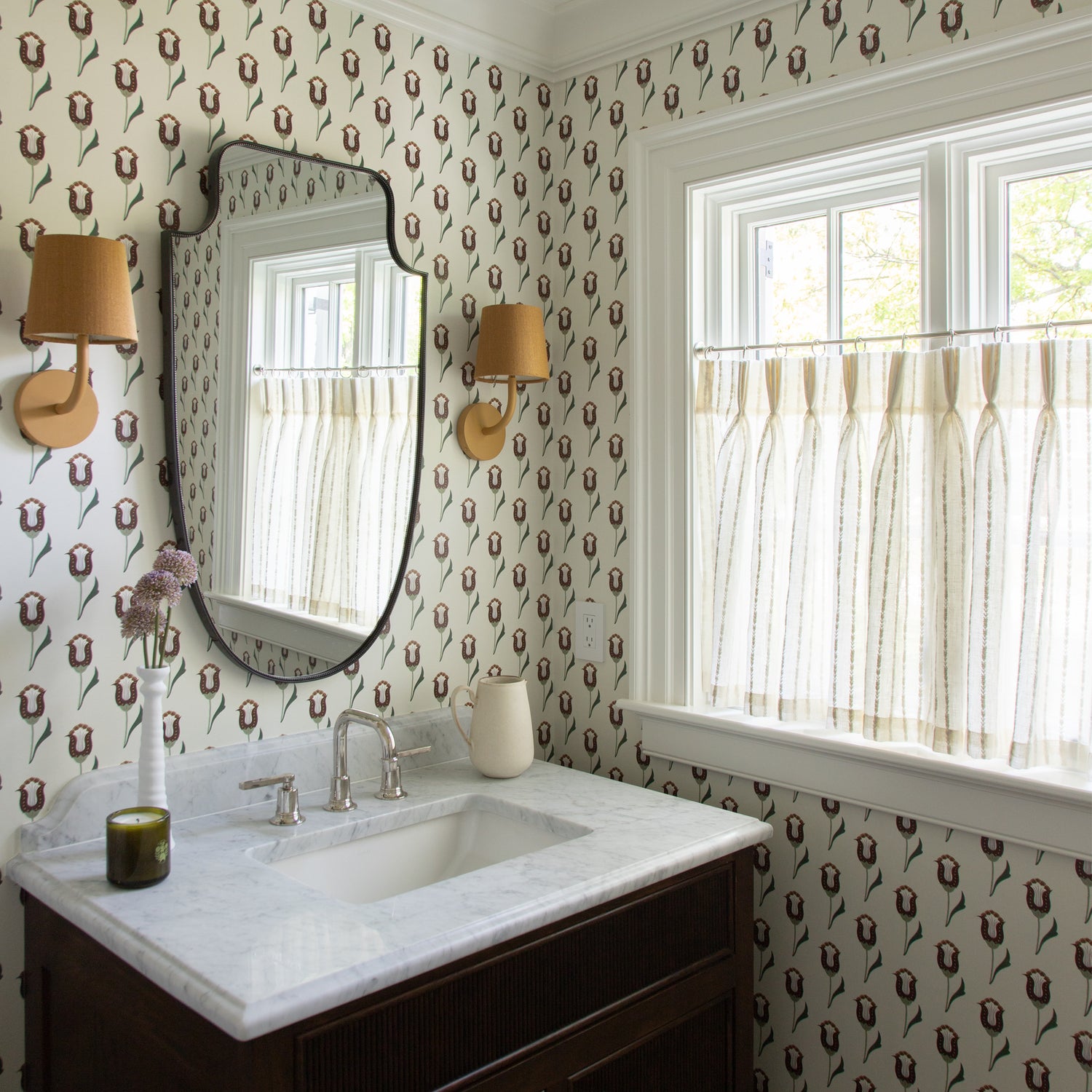 abstract floral pattern wallpaper with neutral background and pink and maroon floral pattern in a bathroom with a mirror hung on the wall and two pale orange lights on either side of the mirror and cream sheer cafe curtains in front of an illuminated window