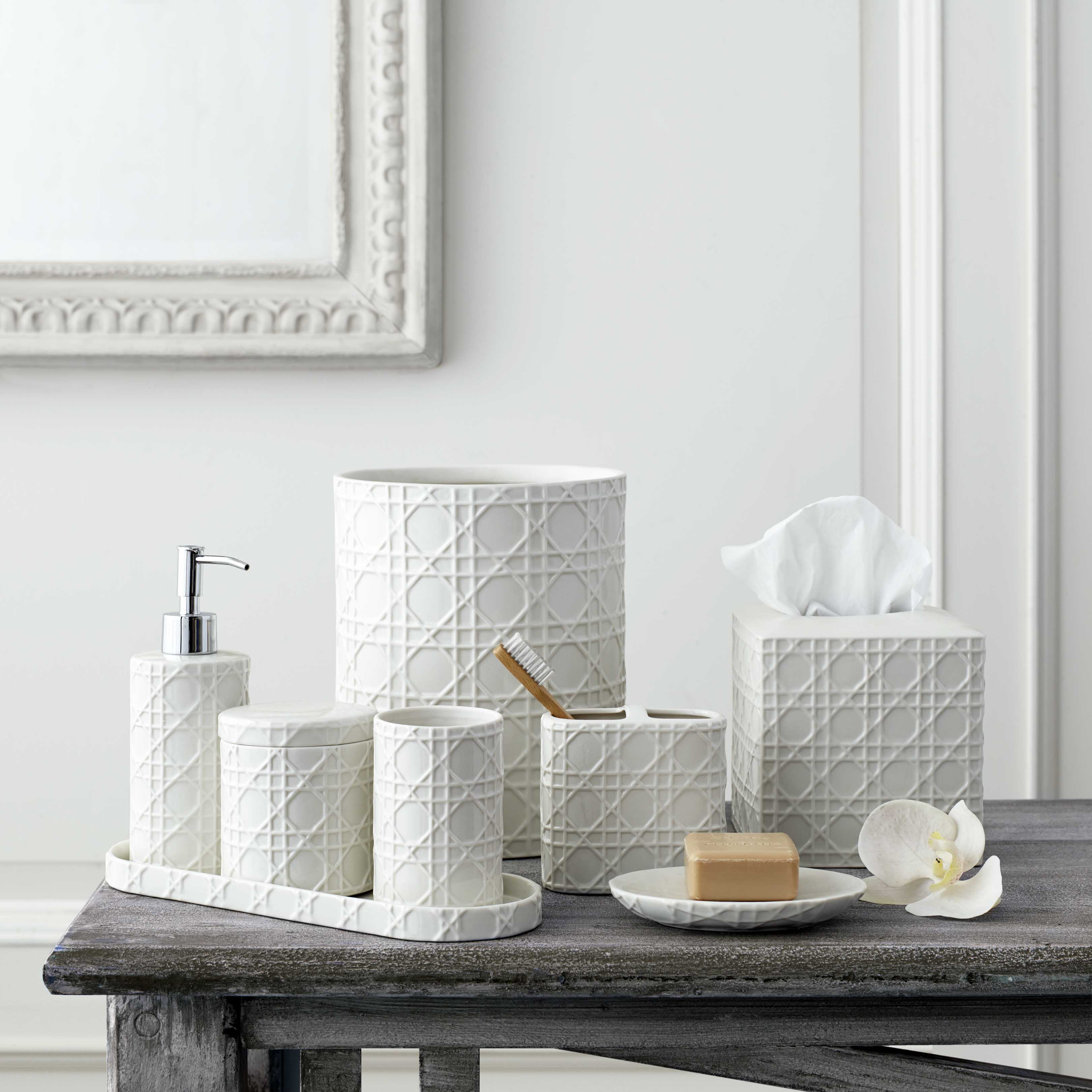 white porcelain rattan accessories tray with tumbler, lotion dispenser, and cotton jar on it, tooth brush holder, waste basket, tissue holder, and soap dish to the right of the tray