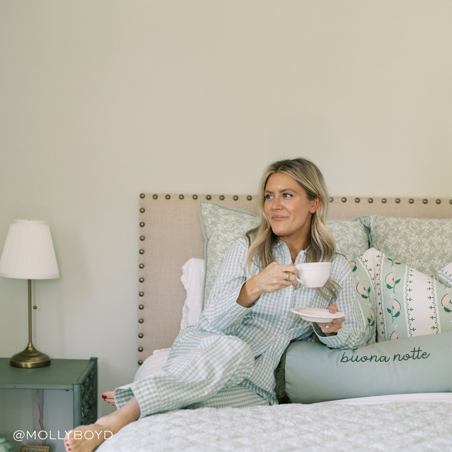 Close-up of bed styled with green and white pillows and sage green monogrammed lumbar with blonde woman wearing pajamas drinking coffee. Photo taken by Molly Boyd