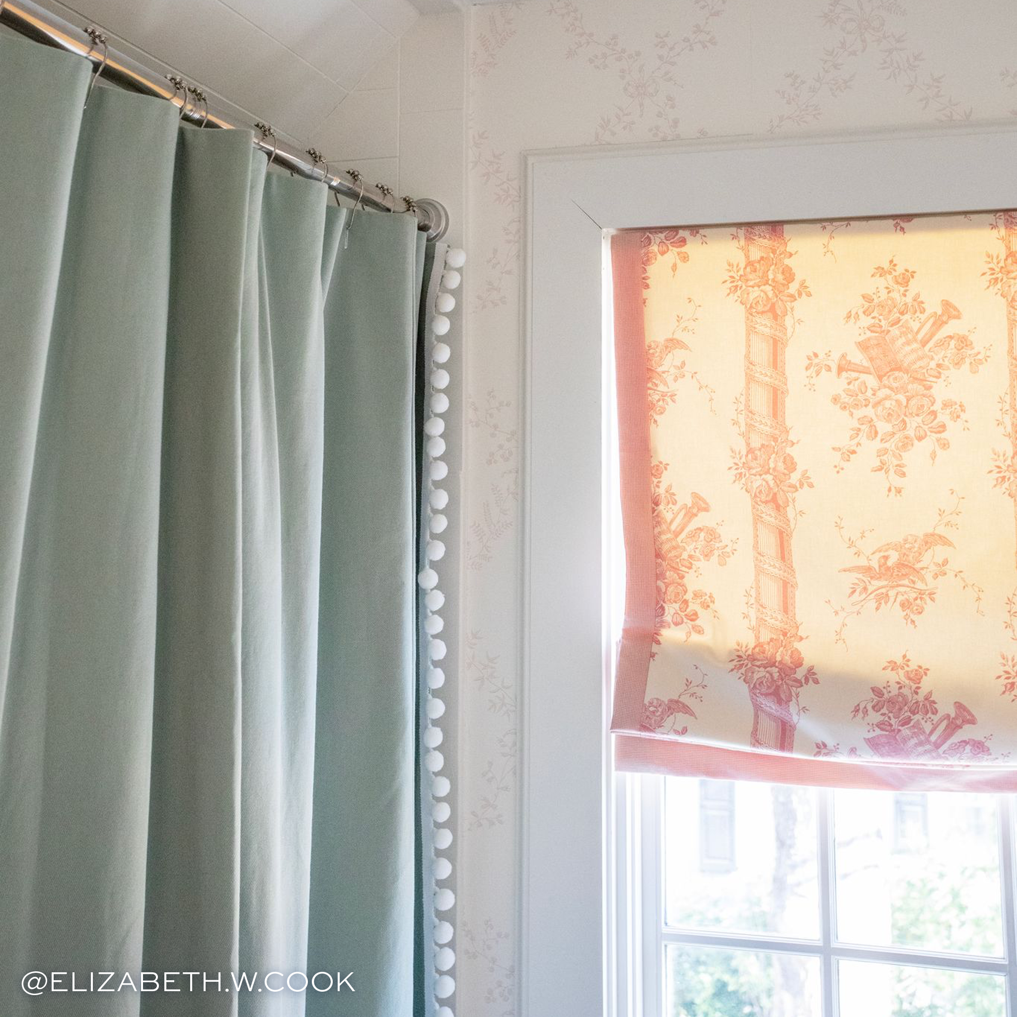 close up of light green cotton shower curtain with white pom pom trim hanging on a rod next to a window with a pink and cream floral shade