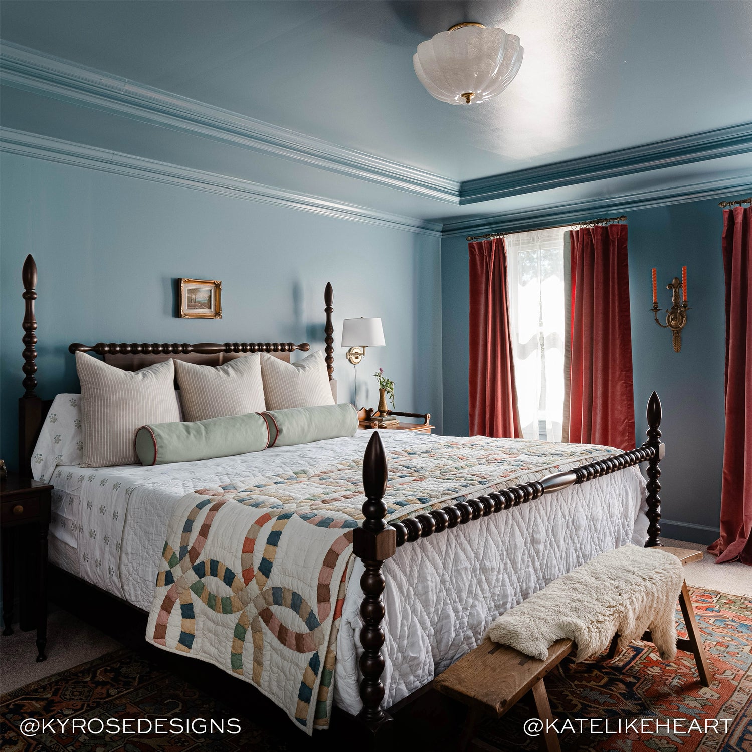 blue bedroom with a brown wooden bed with white bedding and sage green bolster pillows, coral velvet curtains hung in front of illuminated windows and a red multicolored rug under the bed