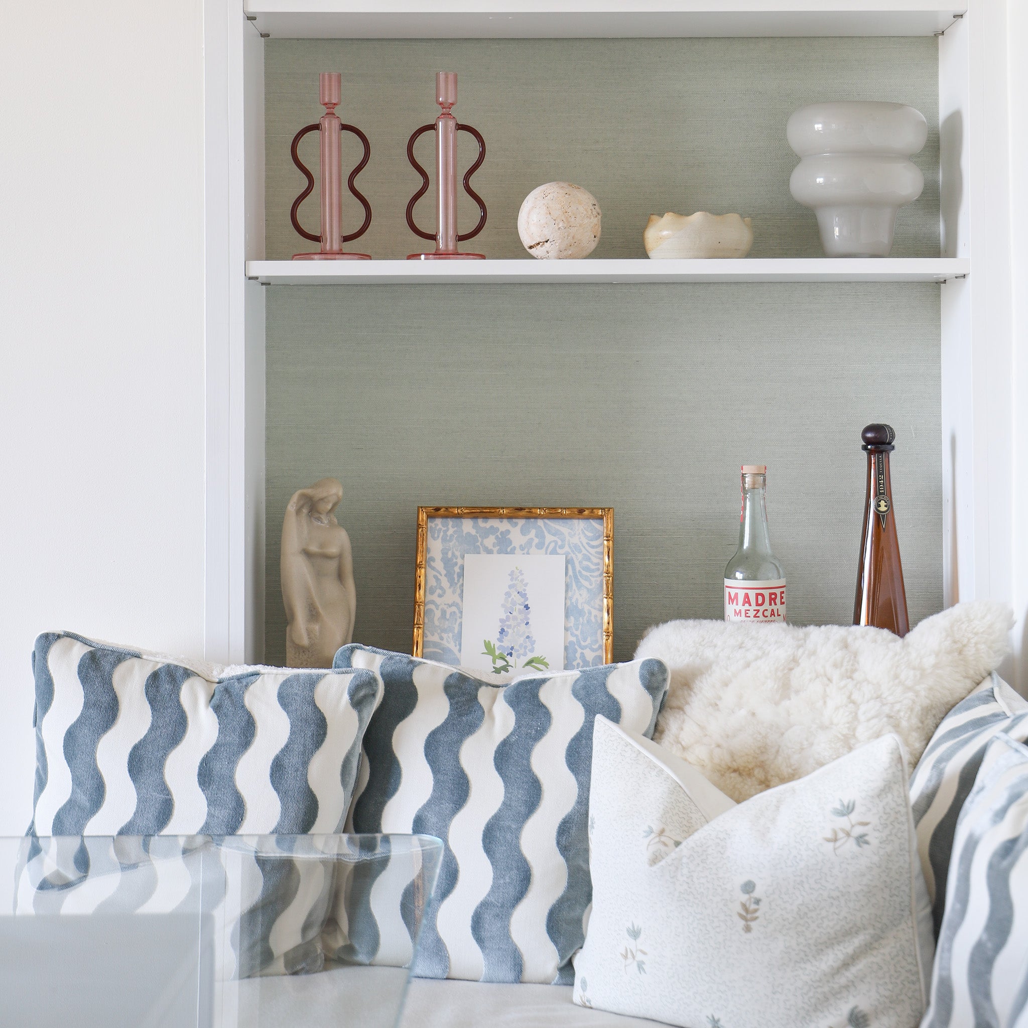 blue green grasscloth wallpaper on the wall with white wooden built in shelves on it with bottles and vases on it and a white couch in front with blue and white pillows on it