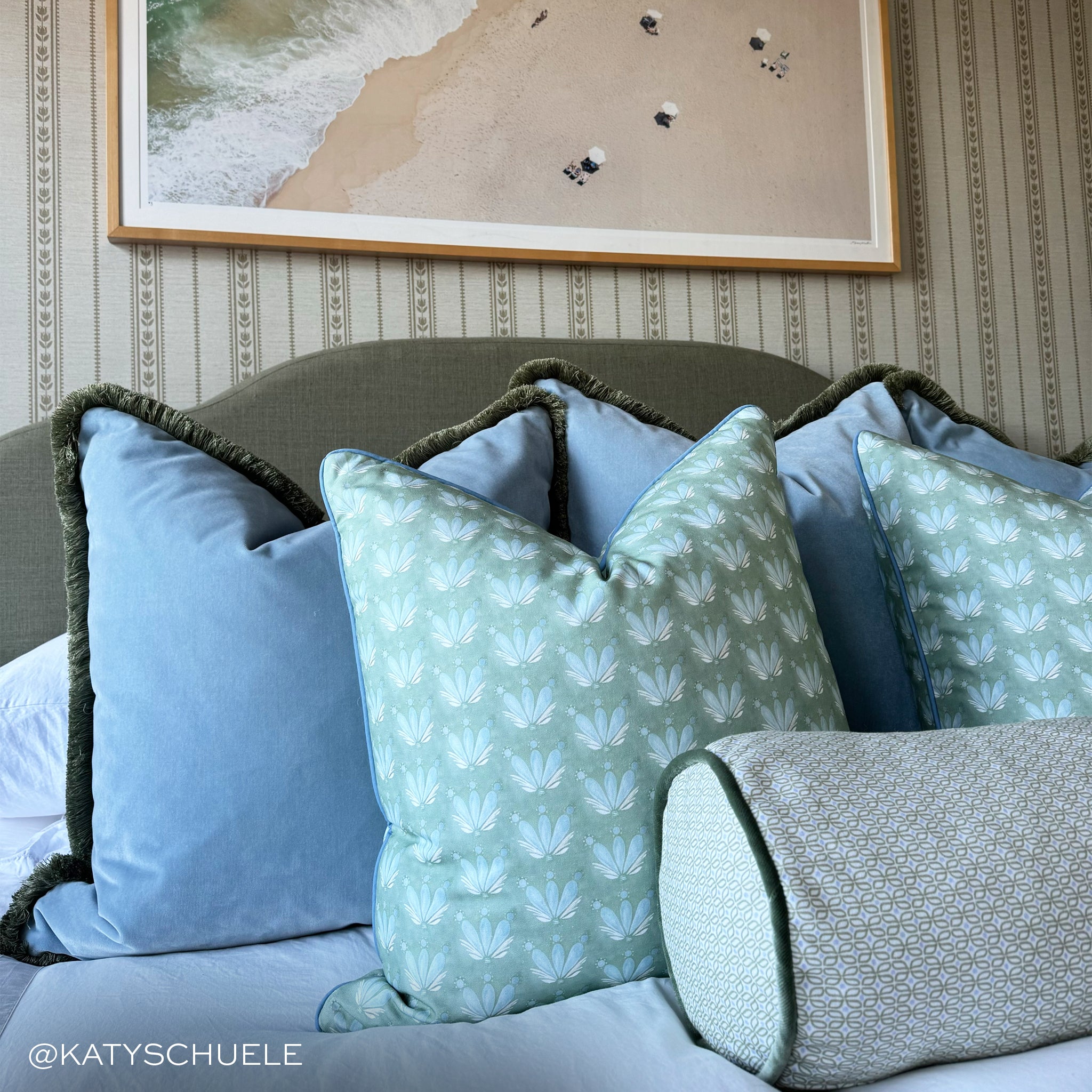 close up of bed with sky blue velvet pillows on it and blue green floral repeat pattern pillows on it with striped wallpaper on the walls