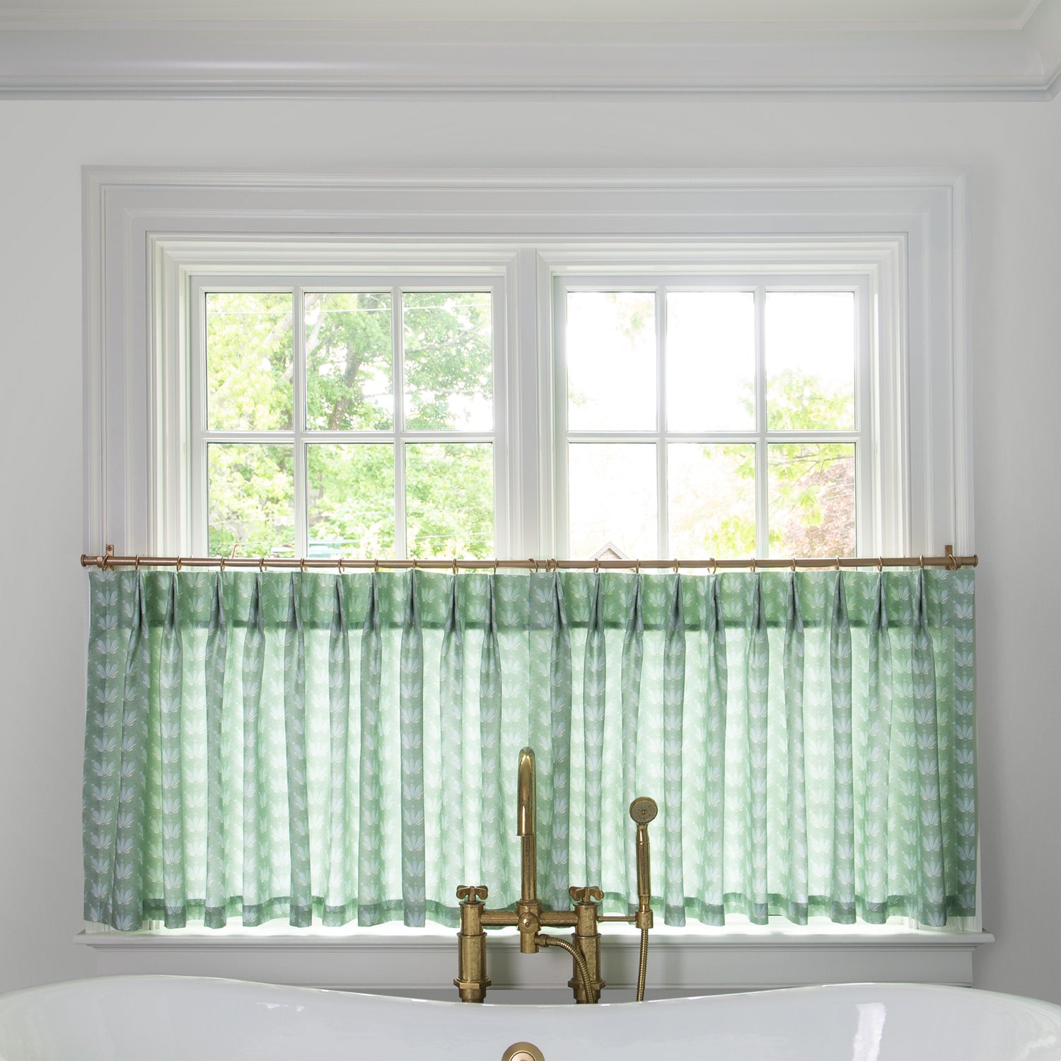 Blue & Green Floral Drop Repeat  cafe curtain hung in front of an illuminated window in a bathroom with a white bathtub in front of the window with a gold faucet 