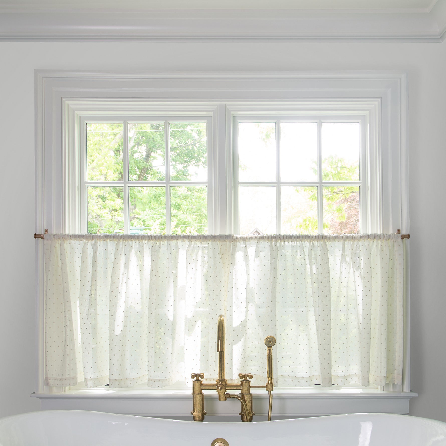 sheer white embroidered cream polka dot cafe curtain hung in front of an illuminated window in a bathroom with a white bathtub in front of the window with a gold faucet 