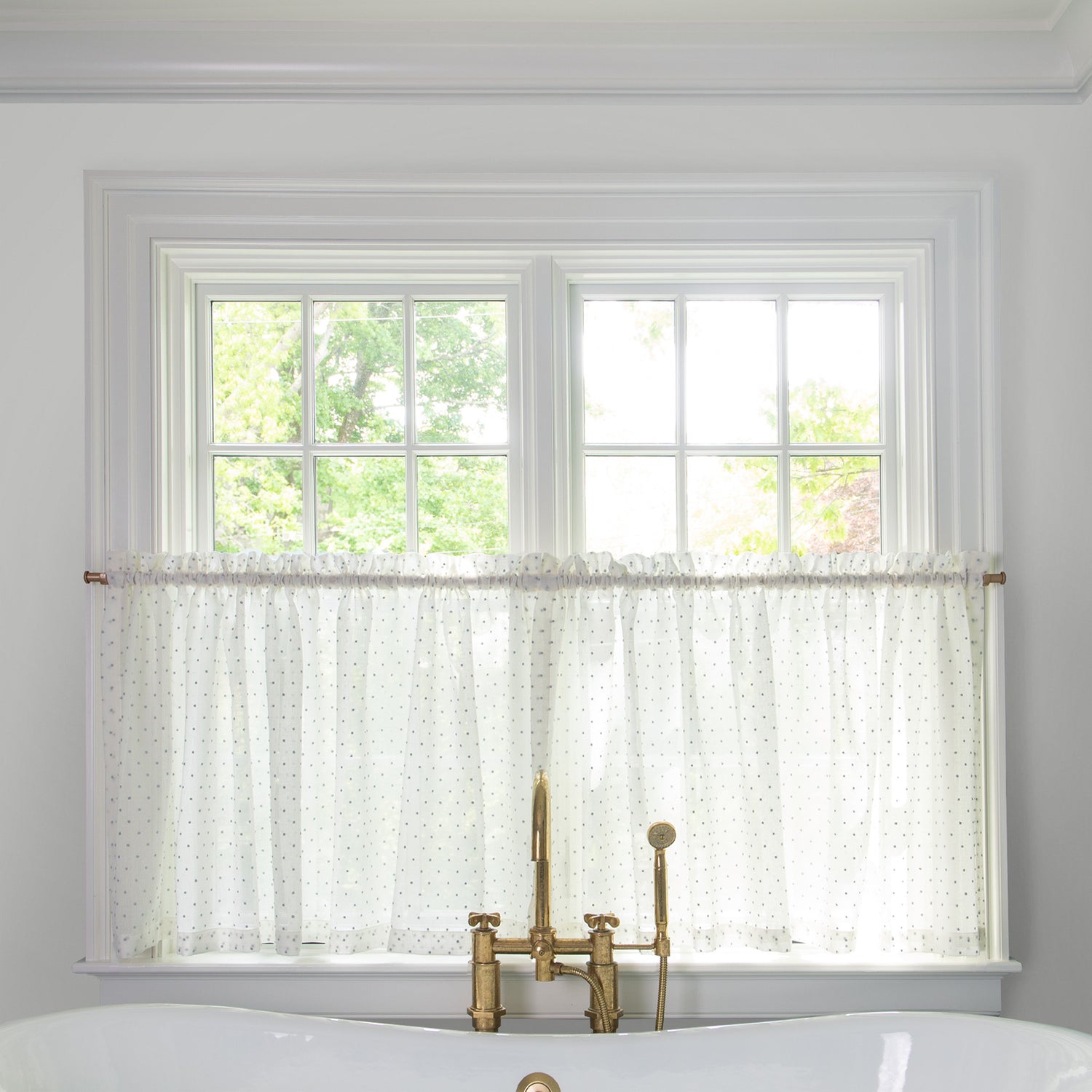 sheer white embroidered blue polka dot cafe curtain hung in front of an illuminated window in a bathroom with a white bathtub in front of the window with a gold faucet 