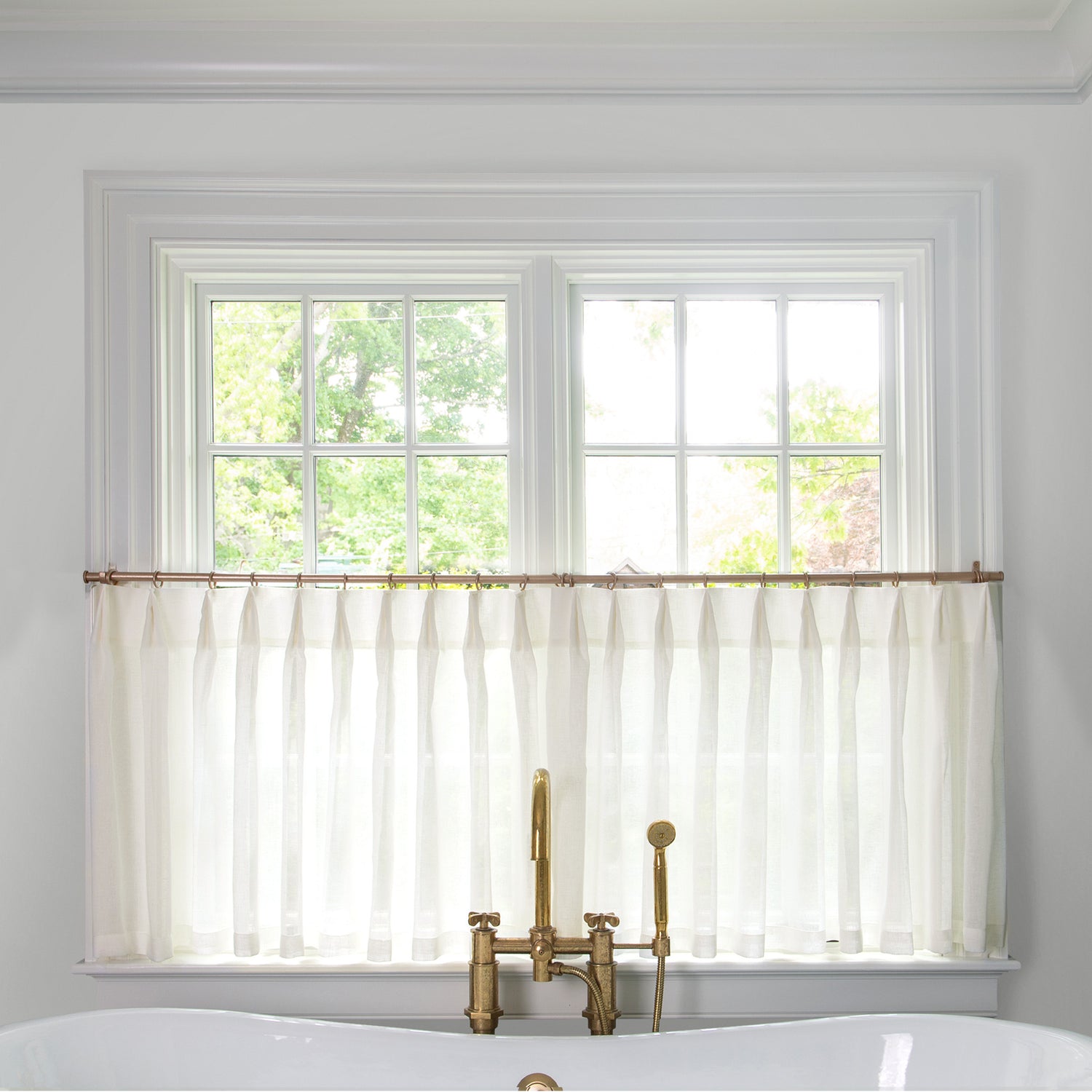 sheer white cafe curtains hung in front of an illuminated window in a bathroom with a white bathtub in front of the window with a gold faucet 