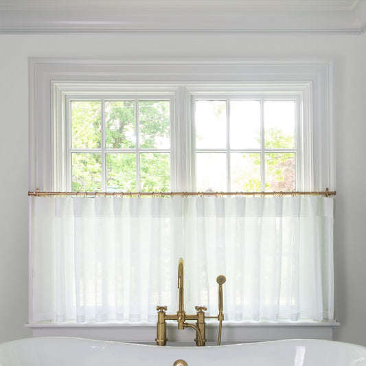 sheer white cafe curtain hung in front of an illuminated window in a bathroom with a white bathtub in front of the window with a gold faucet 