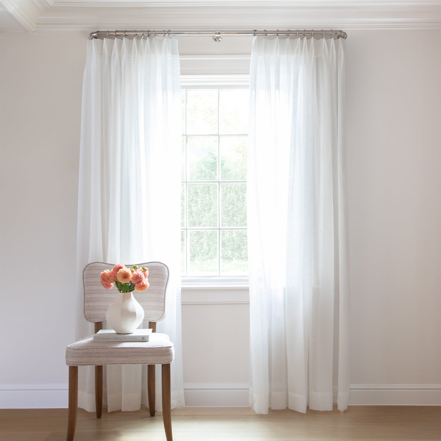 photo of sheer white curtains  hung on a metal rod in front of an illuminated window with a cream chair in front of the window with a cream book and a cream vase with coral flowers in it sitting on the chair