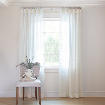 photo of sheer white curtains with embroidered cream polka dots hung on a metal rod in front of an illuminated window with a cream chair in front of the window with a cream book and a white vase with green plants in it sitting on the chair