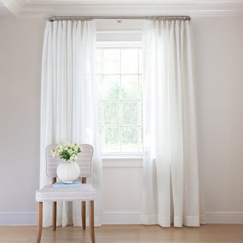 photo of sheer white curtains with embroidered blue polka dots hung on a metal rod in front of an illuminated window with a cream chair in front of the window with a blue book and a white vase with white flowers sitting on the chair