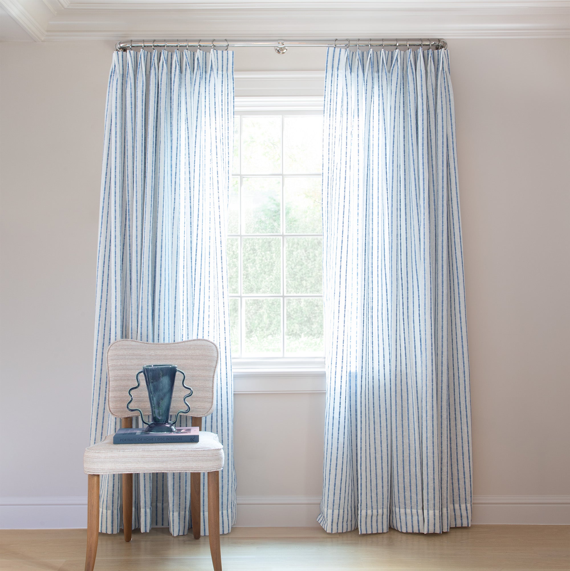 photo of sheer white curtains with embroidered blue stripe pattern hung on a metal rod in front of an illuminated window with a cream chair in front of the window with a blue book and a blue vase sitting on the chair