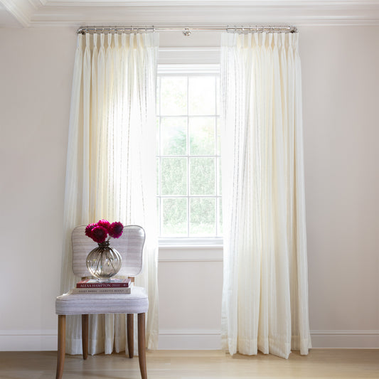 photo of sheer cream curtains with embroidered cream stripe hung on a metal rod in front of an illuminated window with a cream chair in front of the window with a maroon and white books and a clear vase with pink flowers sitting on the chair